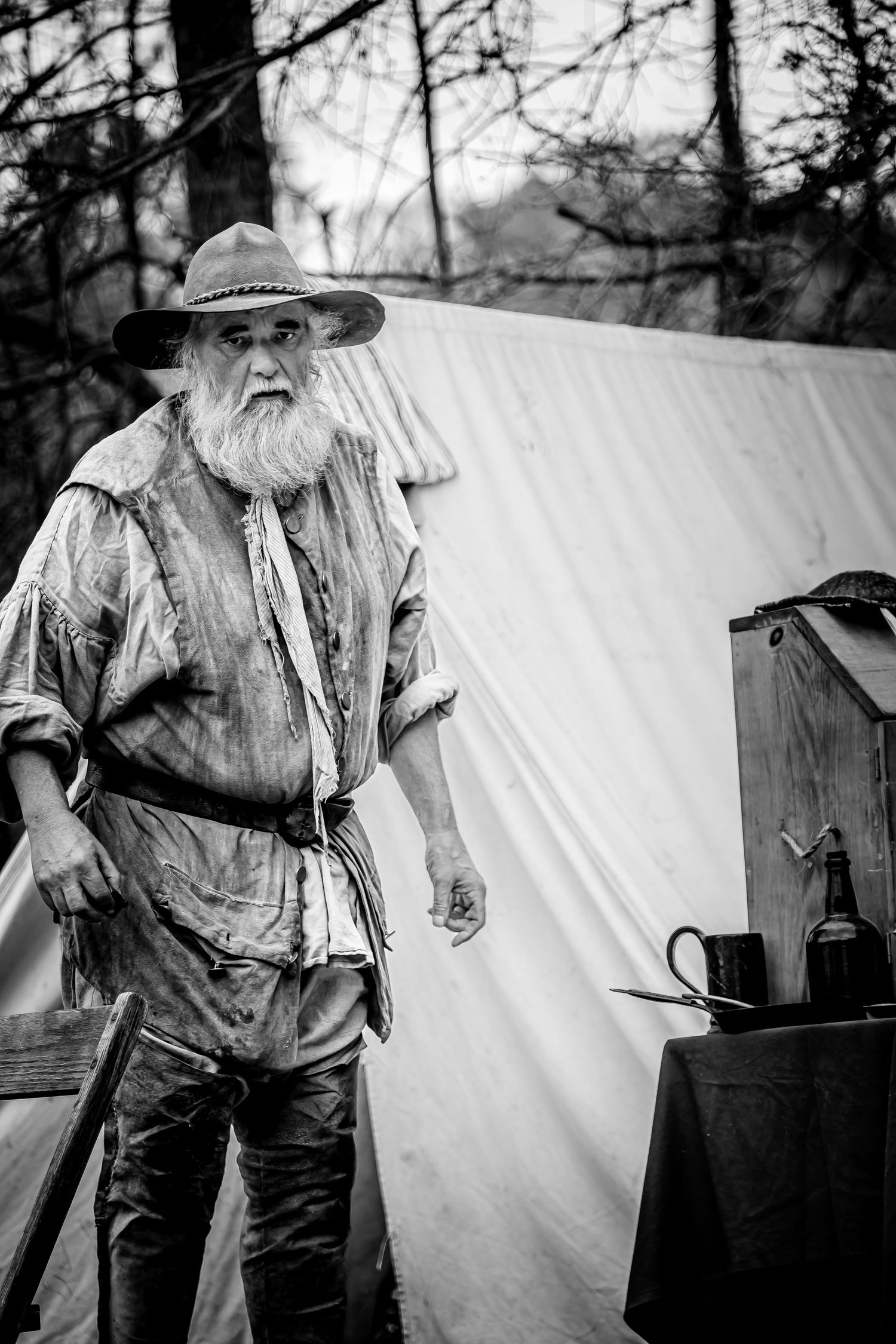 Rugged bearded man in worn frontier buckskin clothing stands by canvas tent, black and white portrait.