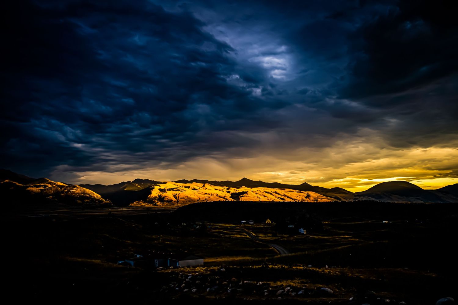 Golden sunset illuminates snow-capped mountains beneath dark stormy skies.