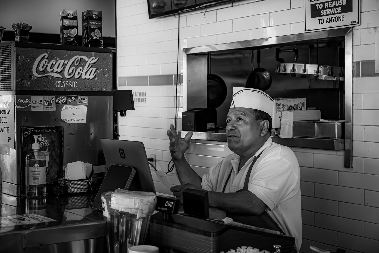 Diner cook in white uniform gestures while standing behind counter with classic Coca-Cola sign in background.