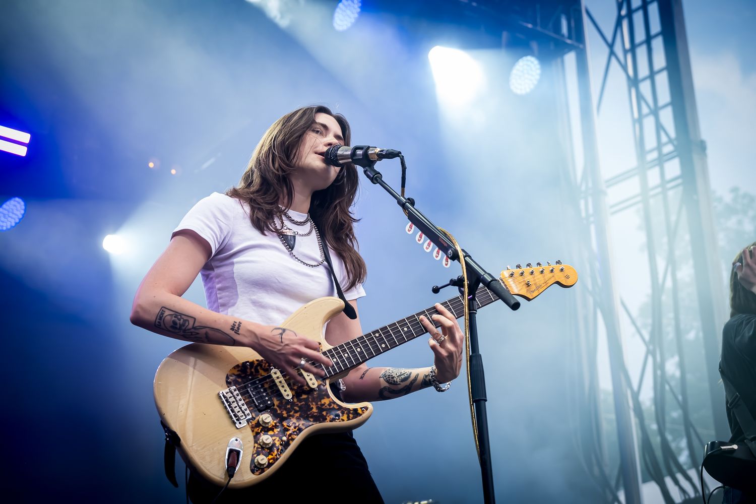 Female guitarist with tattoos playing Fender guitar and singing on stage under bright concert lights.