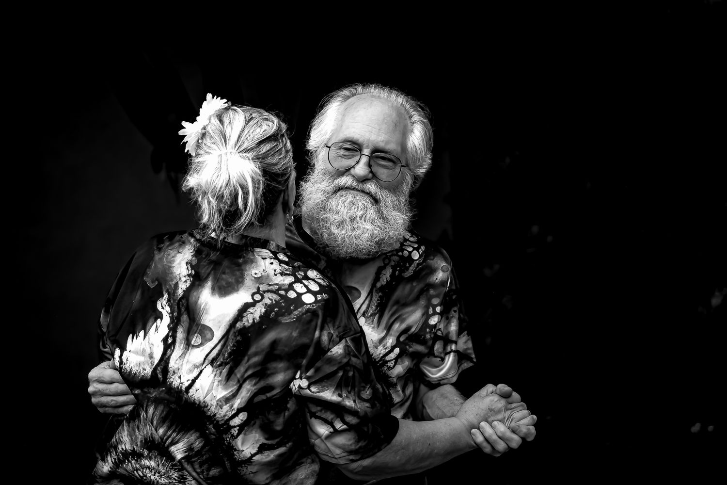 Black and white photo of a smiling elderly bearded man with glasses dancing with a woman in a floral dress.