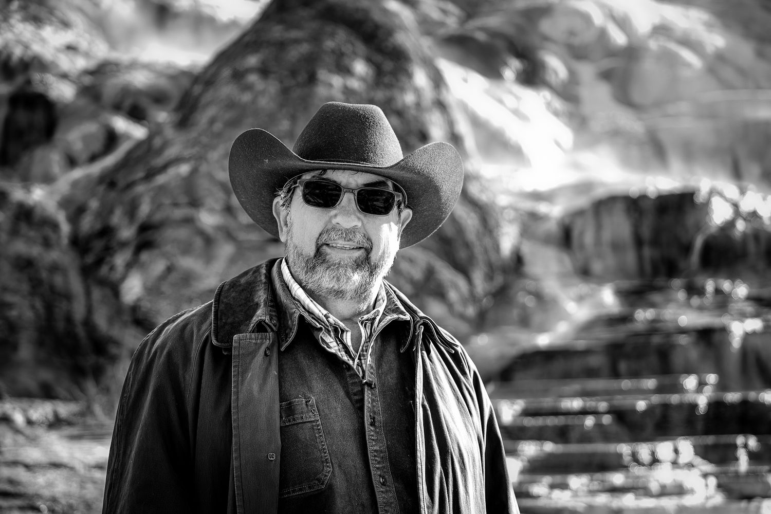 Black and white portrait of a bearded man wearing a cowboy hat and sunglasses with rocky mountains in the background.