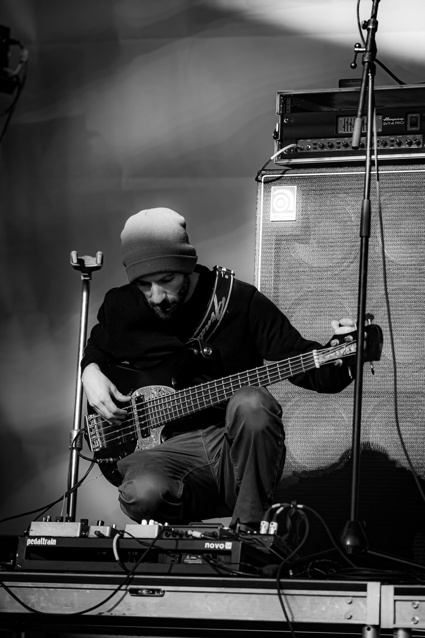 Black and white image of a bassist playing guitar while seated near amplifiers and effect pedals on stage.