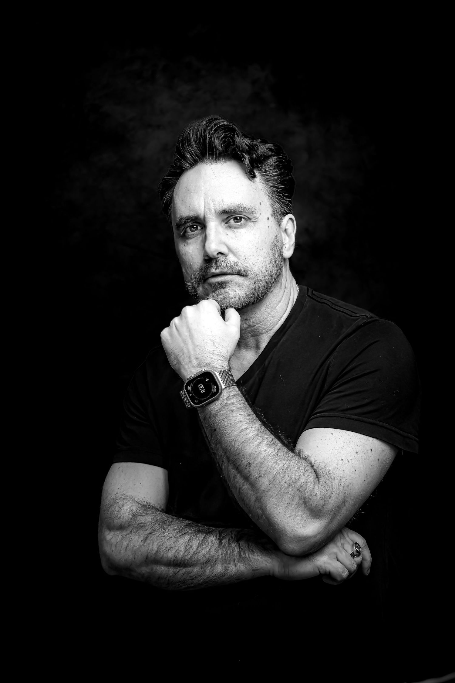 Black and white portrait of a man in a black t-shirt, arms crossed, resting chin on fist, wearing a smartwatch.