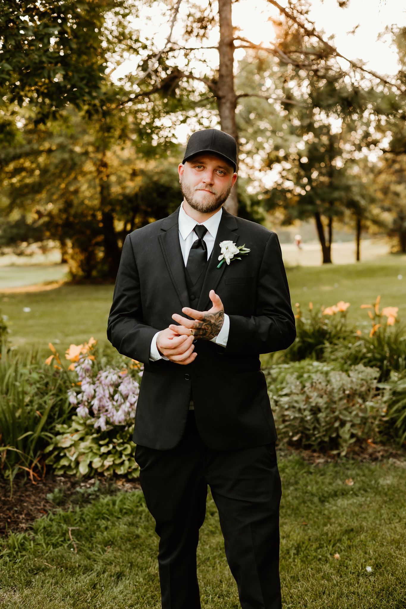 A groom in a black suit with boutonniere and baseball cap stands in a garden setting.