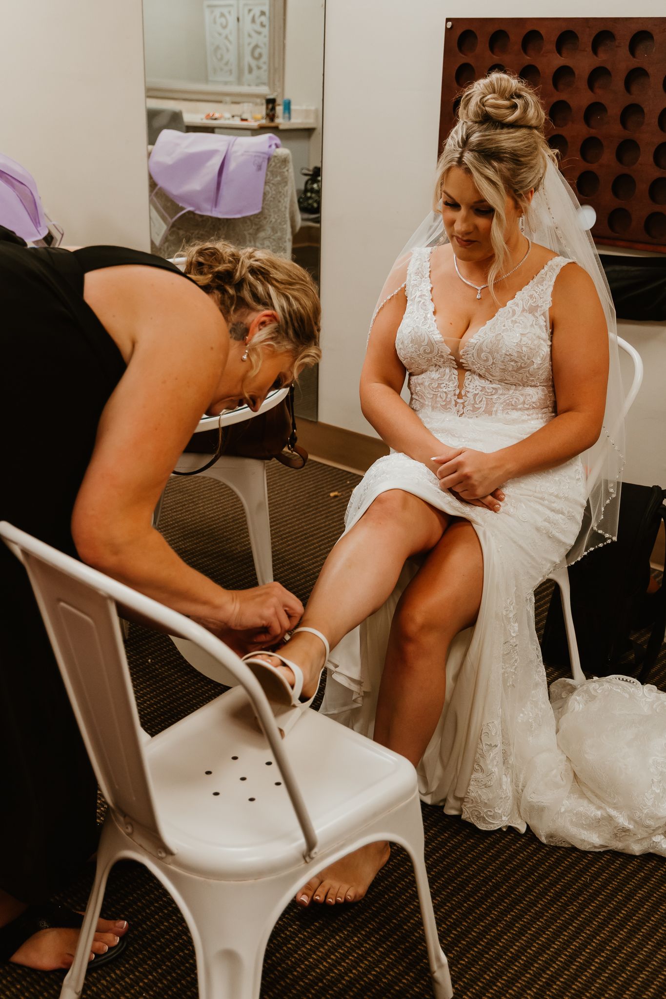 Bride in lace wedding dress sits as bridesmaid helps fasten white heels during wedding preparations.