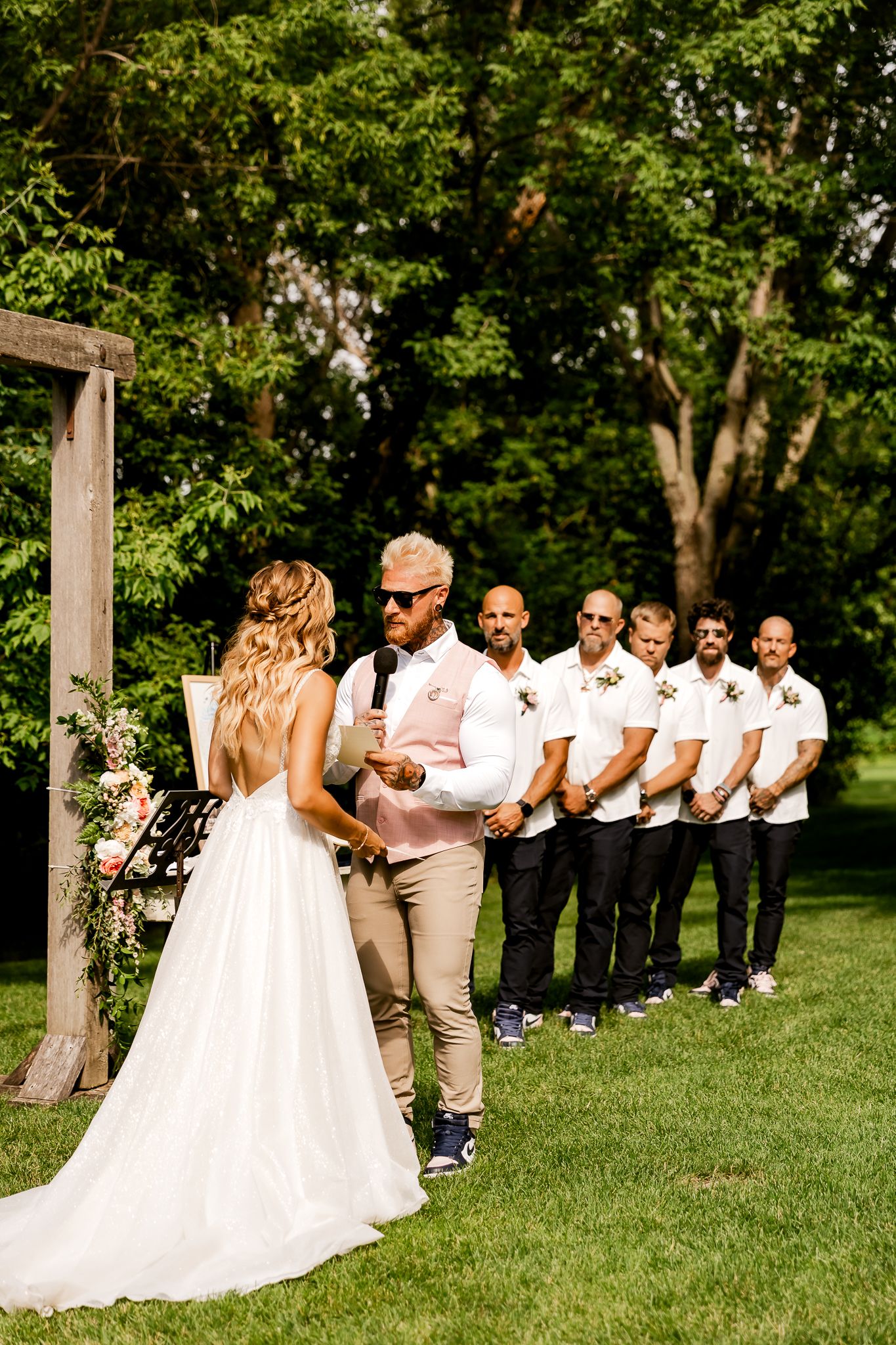 Outdoor wedding ceremony with bride in white gown facing groom in pink shirt, groomsmen in white shirts standing beside wooden arch.
