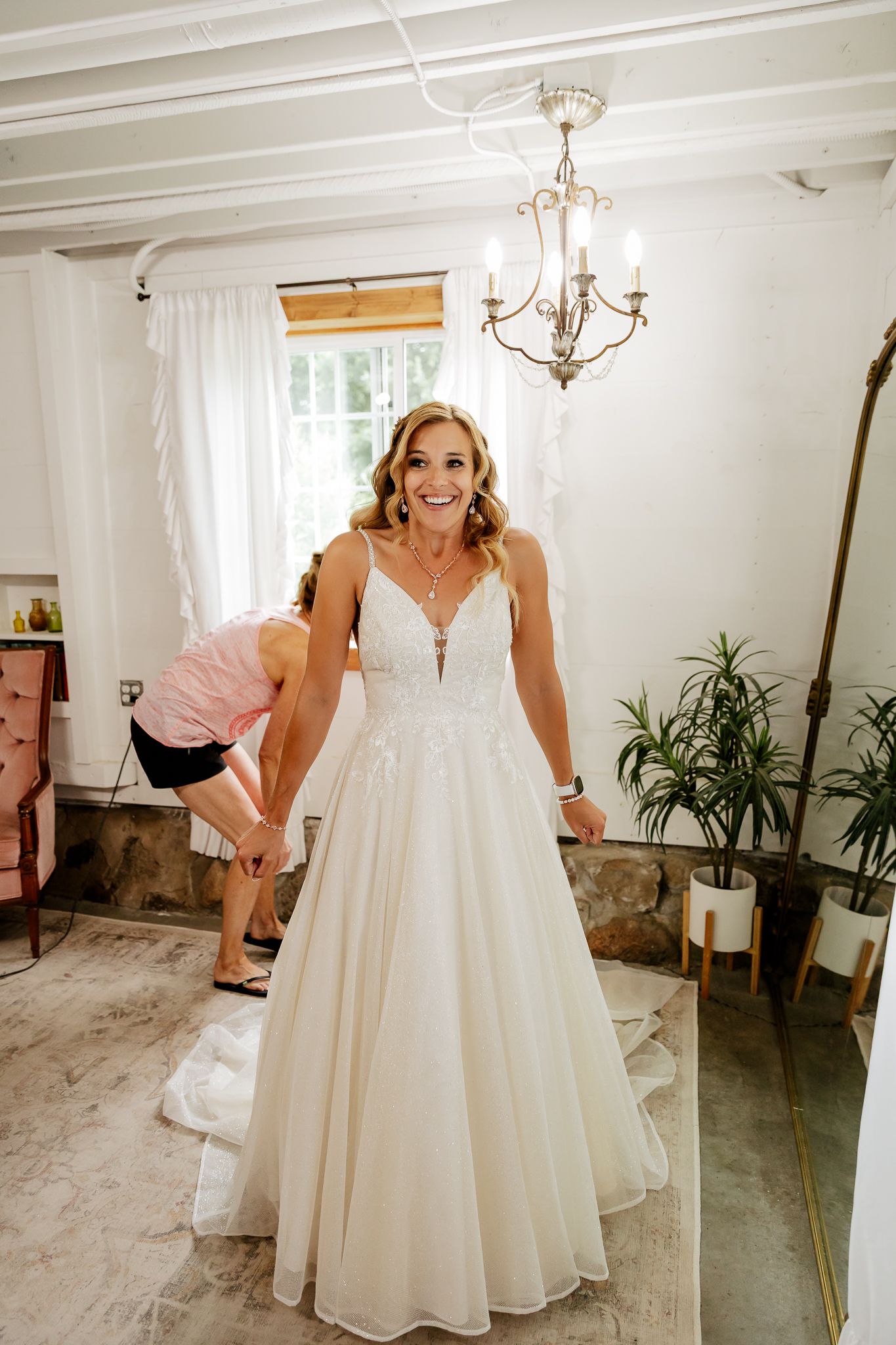 Smiling bride in a white lace A-line wedding gown being helped by a bridesmaid in an elegant room.