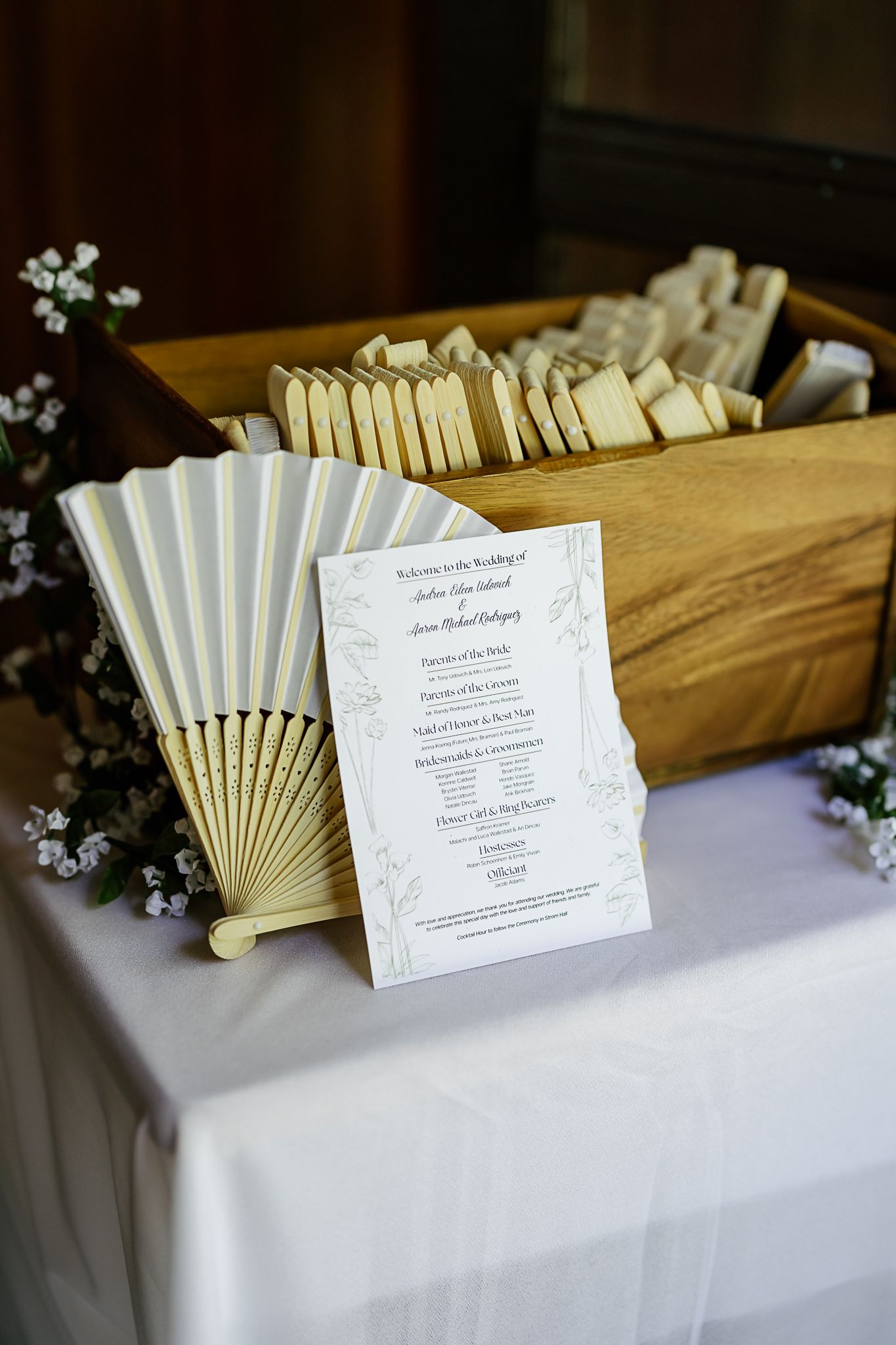 Elegant wedding program display with white hand fans in a wooden box, decorated with delicate white flowers on a draped table.