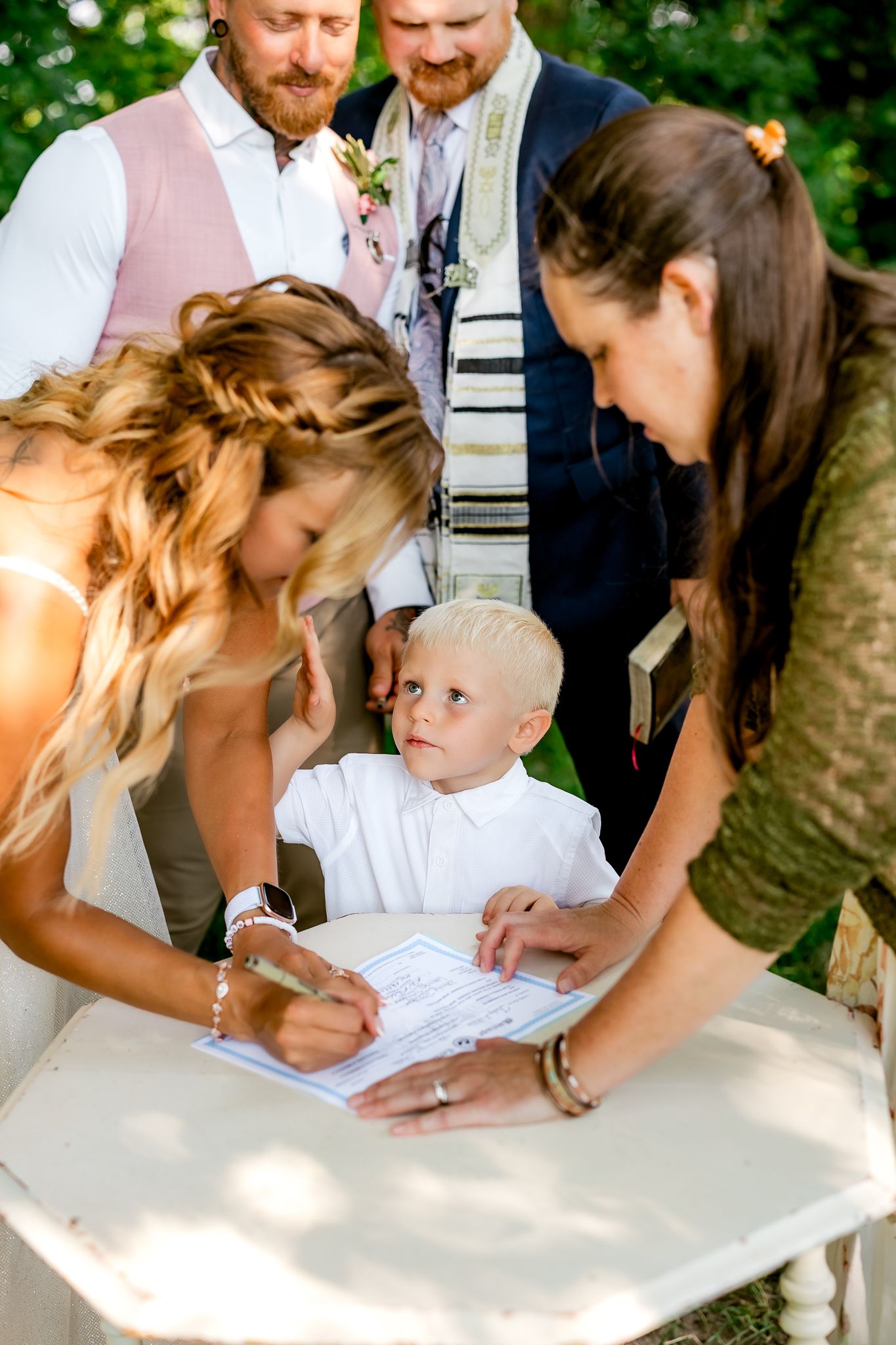 Bride signing wedding documents at outdoor ceremony with young boy watching and guests standing behind.