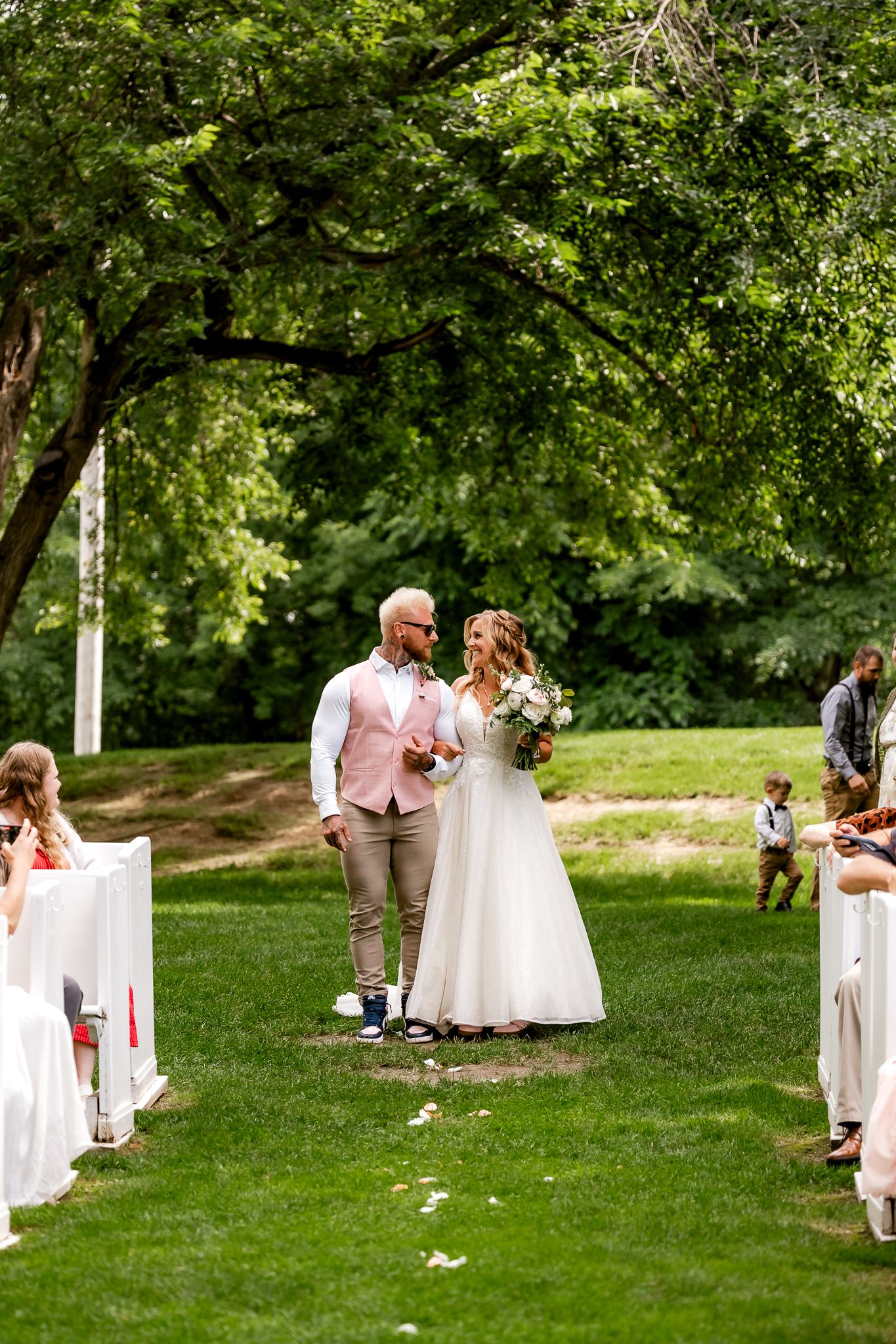 Bride and groom walk down outdoor aisle surrounded by lush green trees and seated guests on a sunny wedding day.
