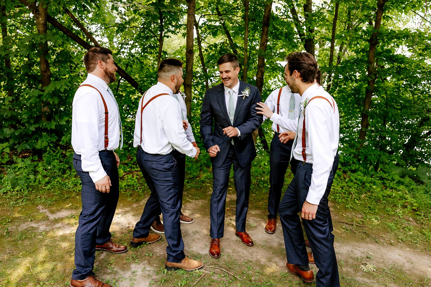 Groom in navy suit with groomsmen in white shirts and suspenders laughing outdoors at a wedding.