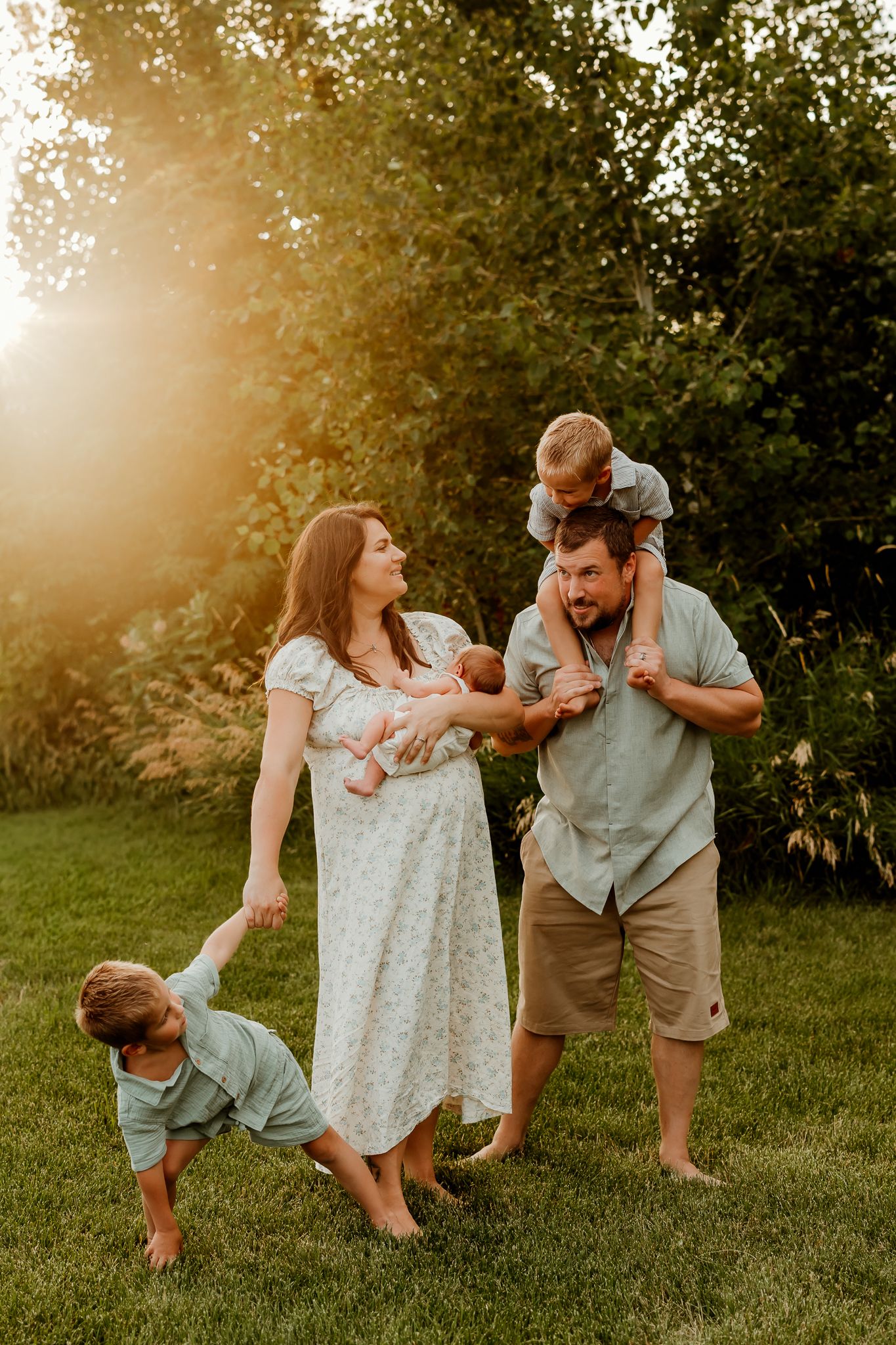 Family of five enjoying golden hour outdoors, mother holding newborn, father with toddler on shoulders, child playing on grass.