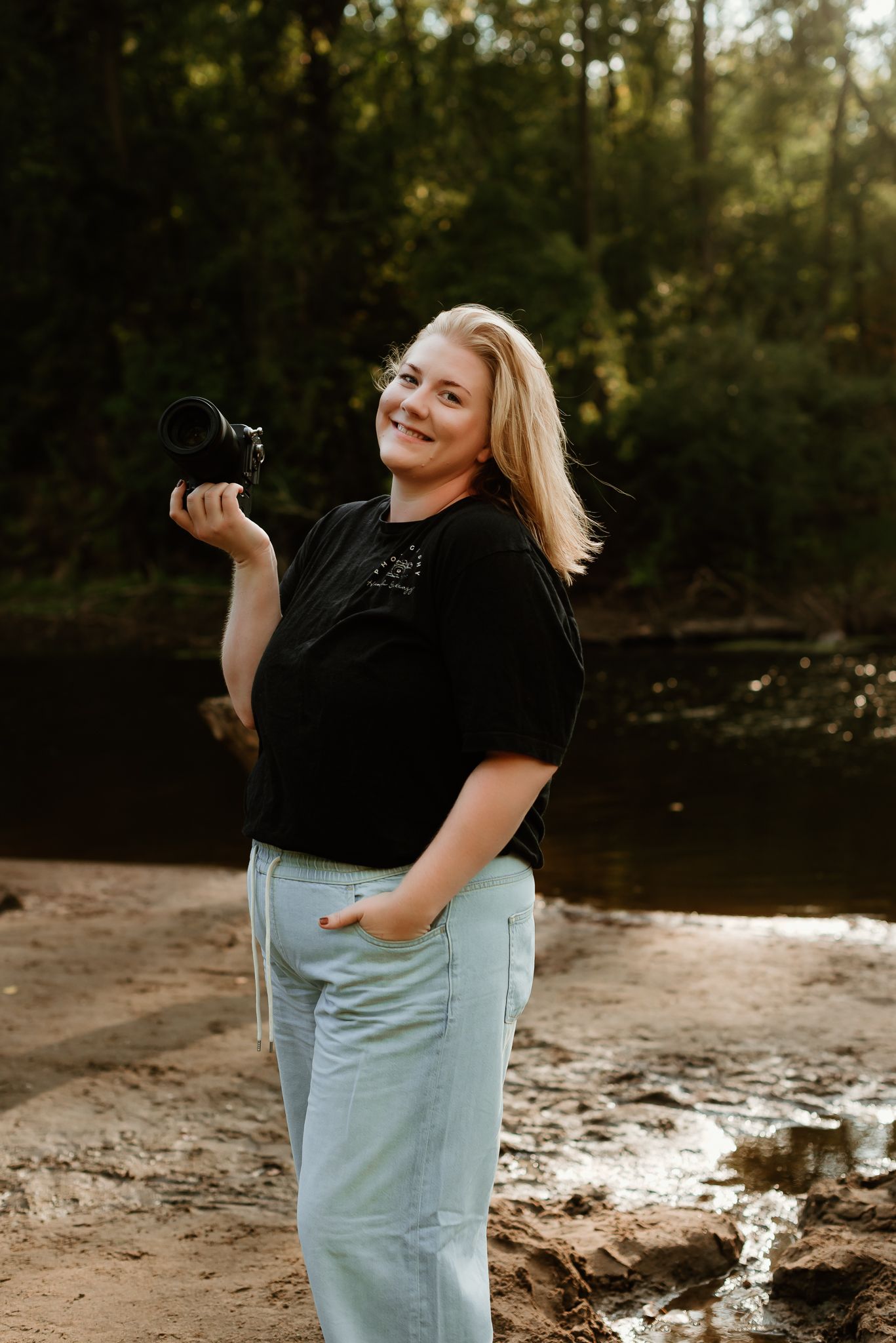 Smiling blonde woman holding a DSLR camera outdoors near a creek, wearing black tee and light jeans.