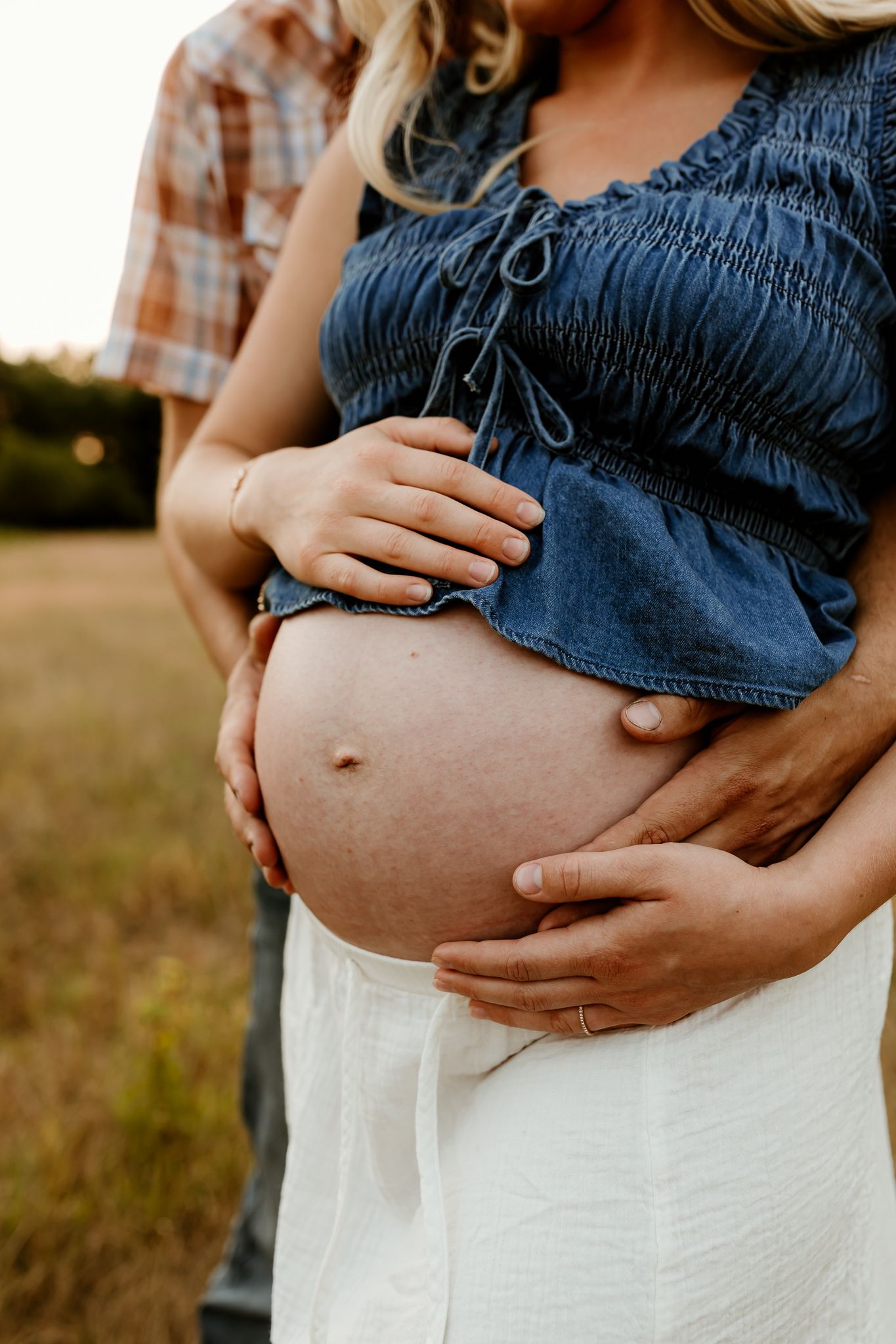 Pregnant woman in denim top and white skirt cradling baby bump outdoors with partner embracing her from behind.