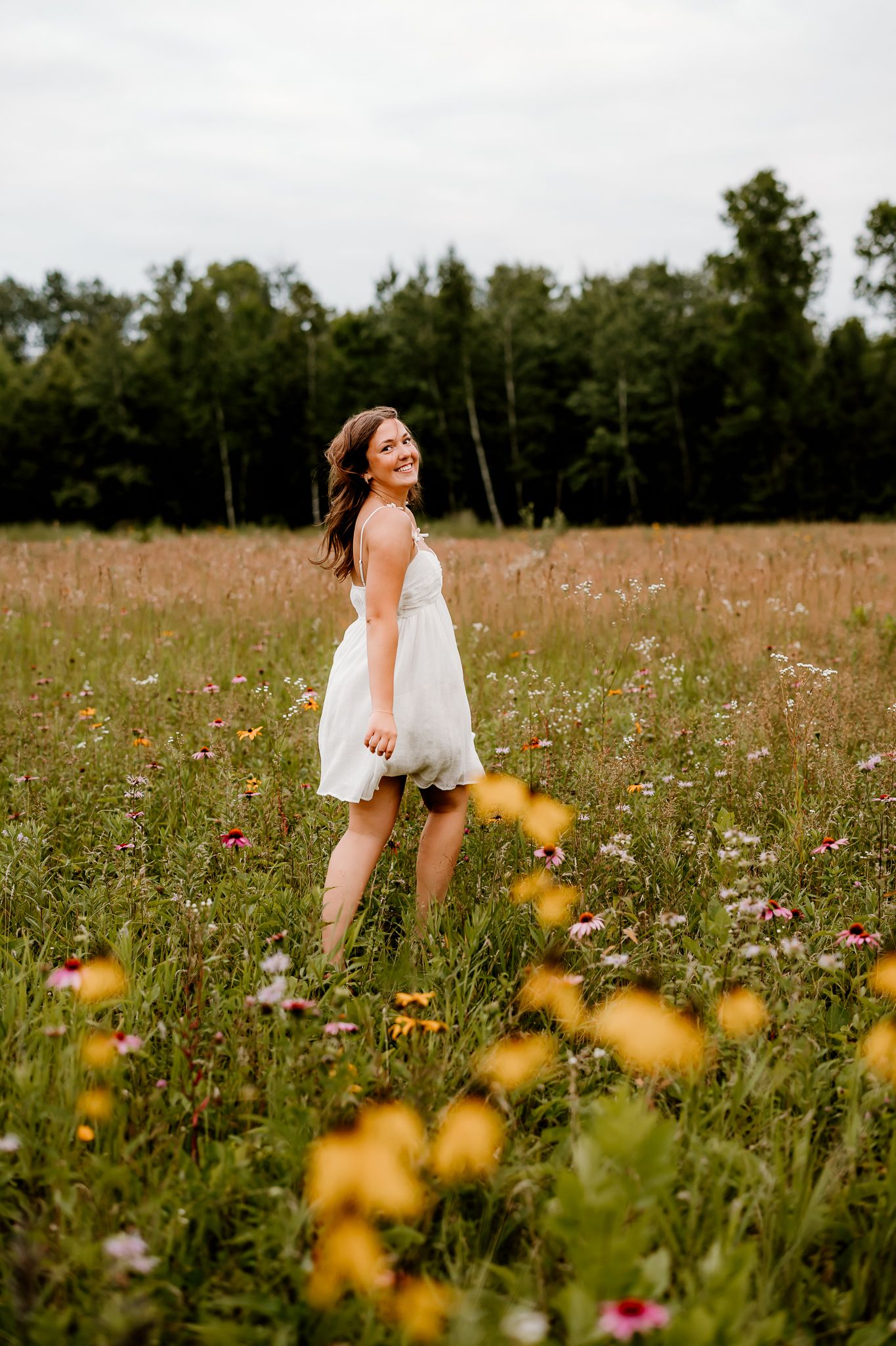 A smiling woman in a white dress walks through a vibrant wildflower meadow with trees in the background.