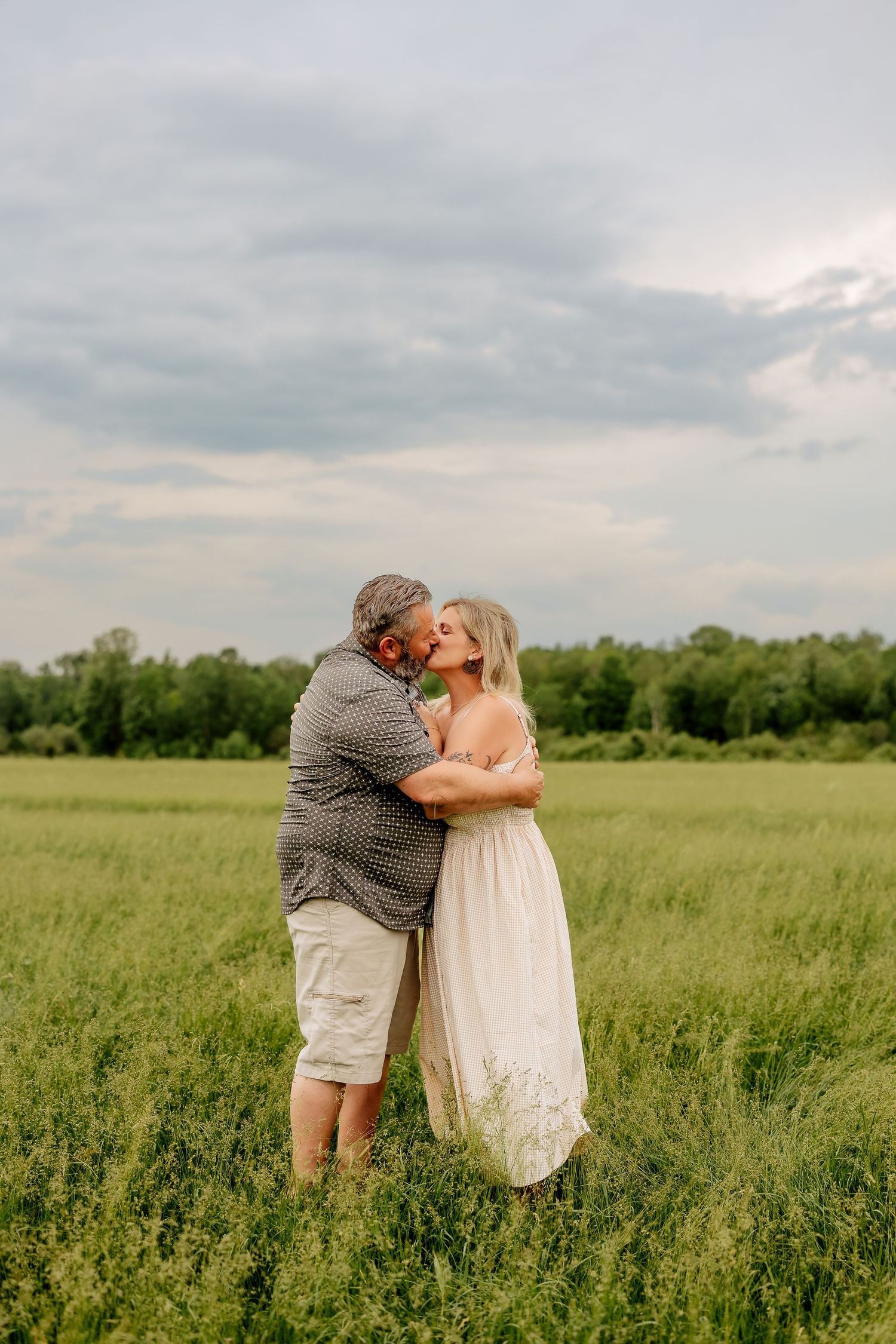 Couple sharing a romantic kiss in a lush green field under cloudy skies.