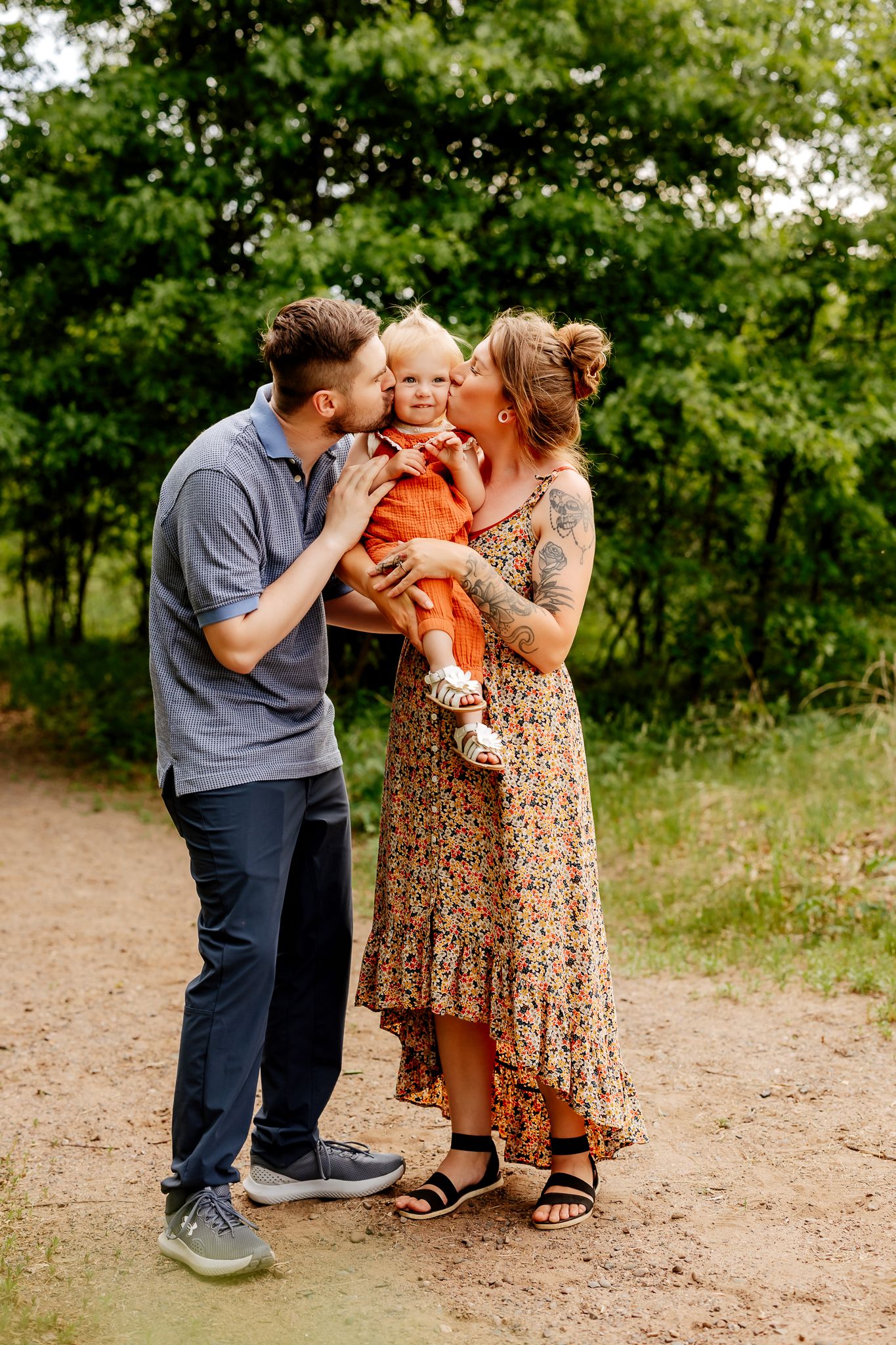 A young family shares kisses with their toddler in an orange outfit on a scenic dirt path surrounded by greenery.
