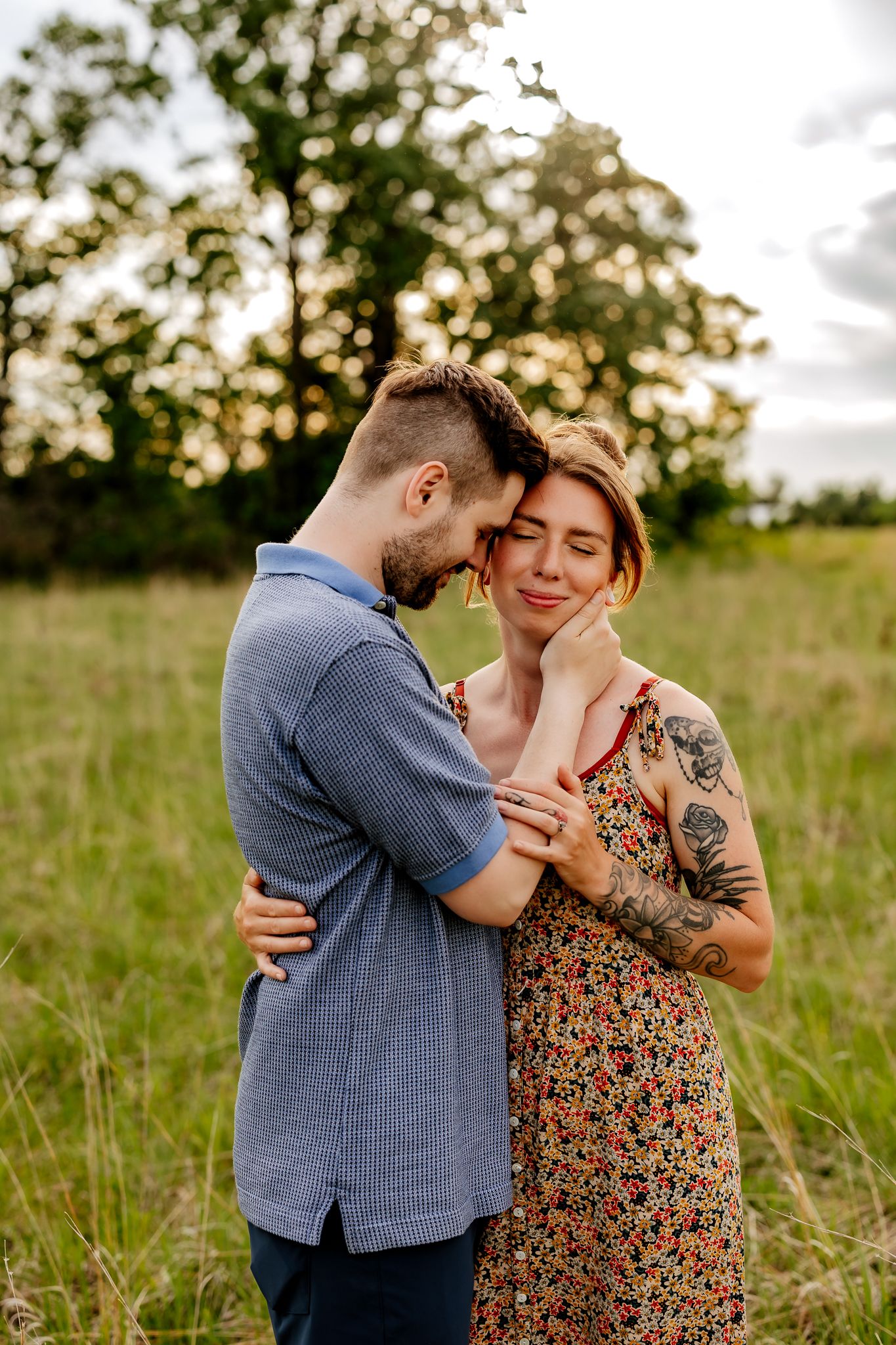 A couple shares a tender embrace in a sunlit field, the woman in a floral dress with visible tattoos.