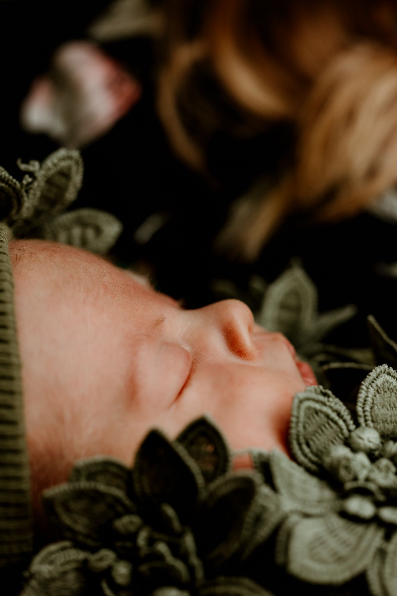 Sleeping newborn baby wrapped in green floral lace fabric with soft bokeh background.
