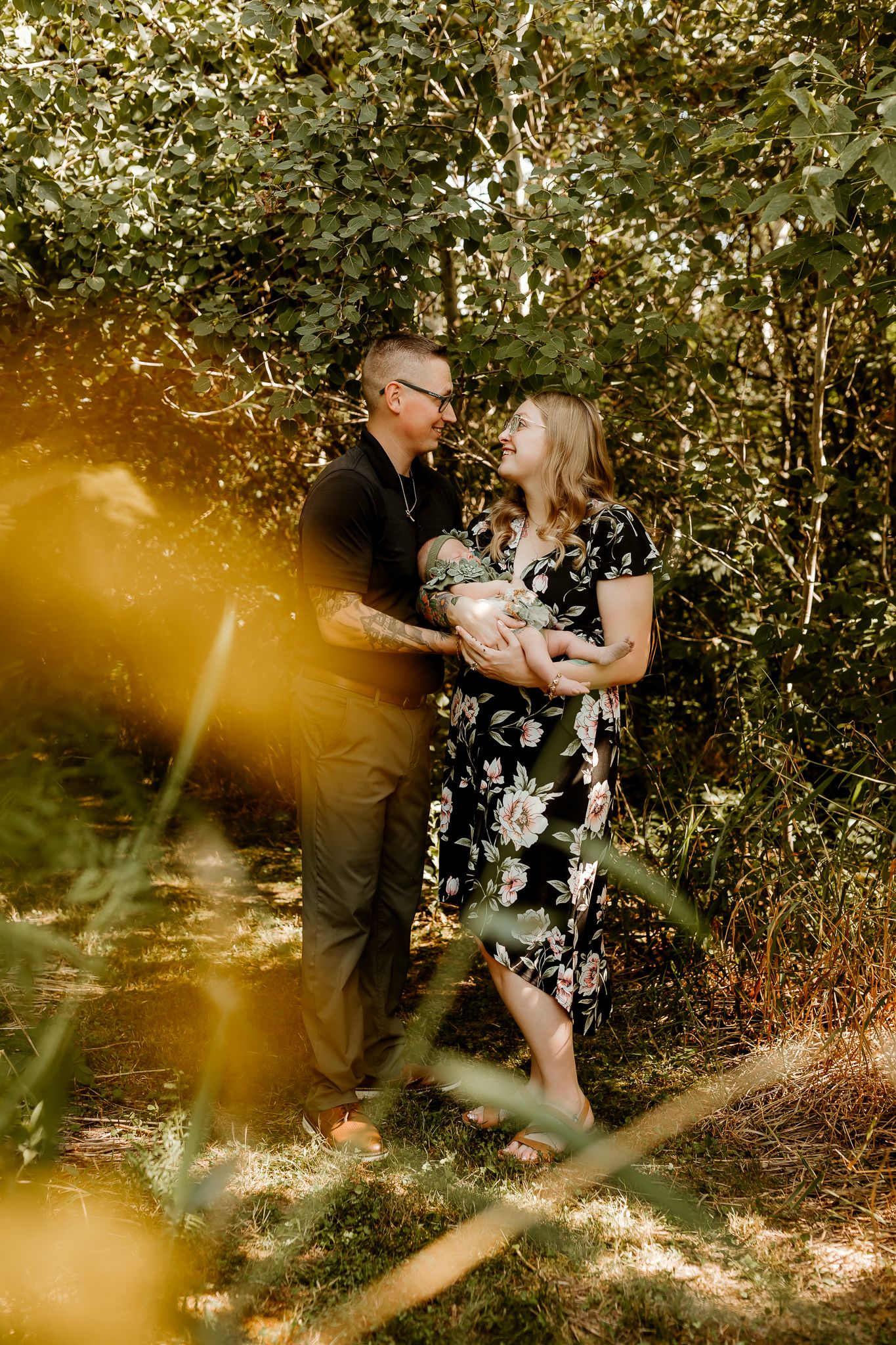 Couple standing in lush green forest, woman in floral dress holding newborn baby, man in black shirt gazing lovingly at her.