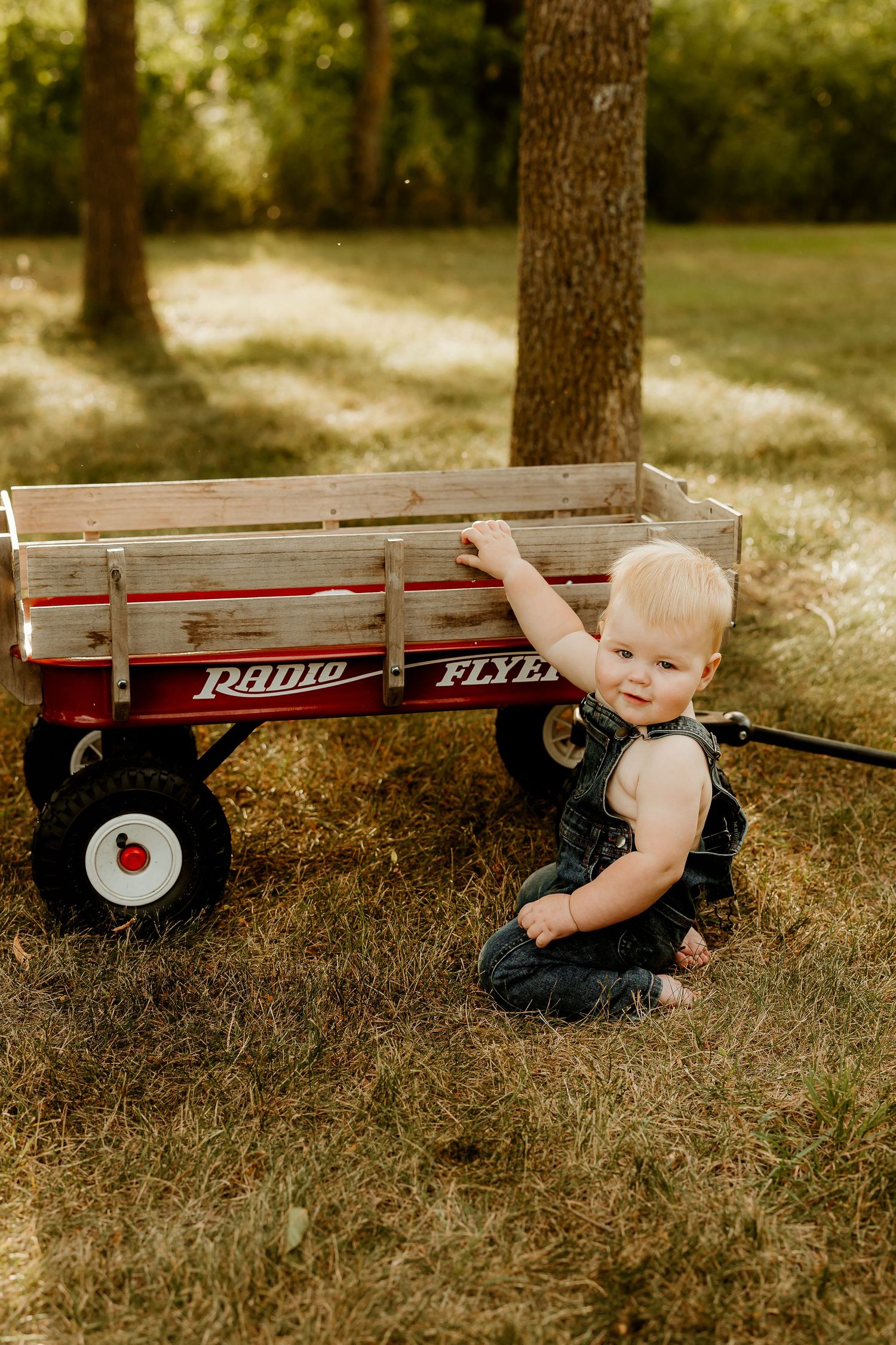 Smiling baby in overalls sitting on grass next to a classic red Radio Flyer wagon outdoors.