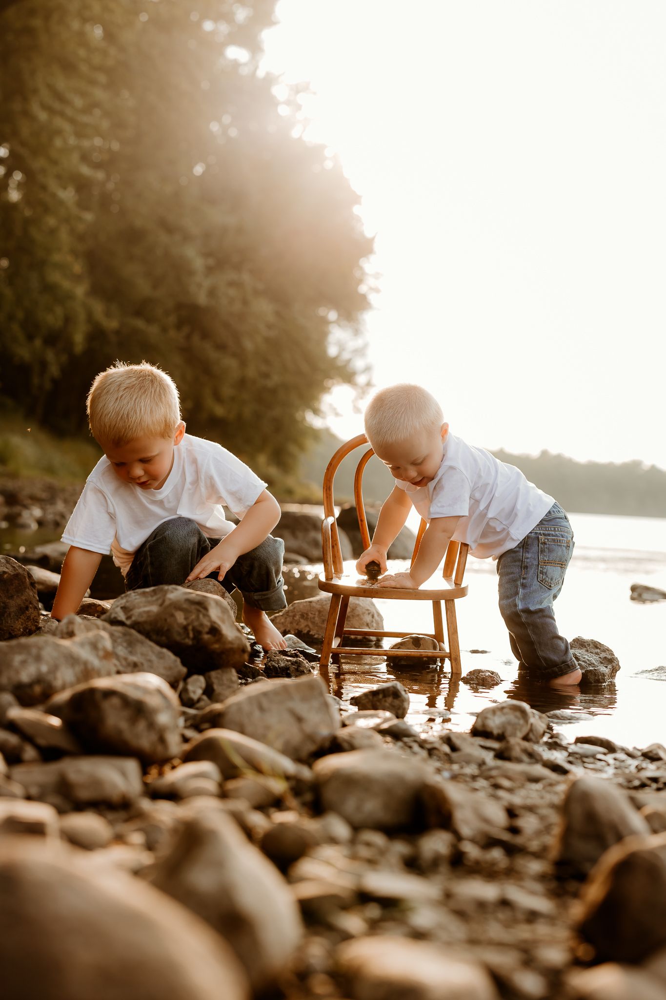Two young boys in white shirts play on a rocky riverbank at golden sunset, exploring stones near a wooden chair.