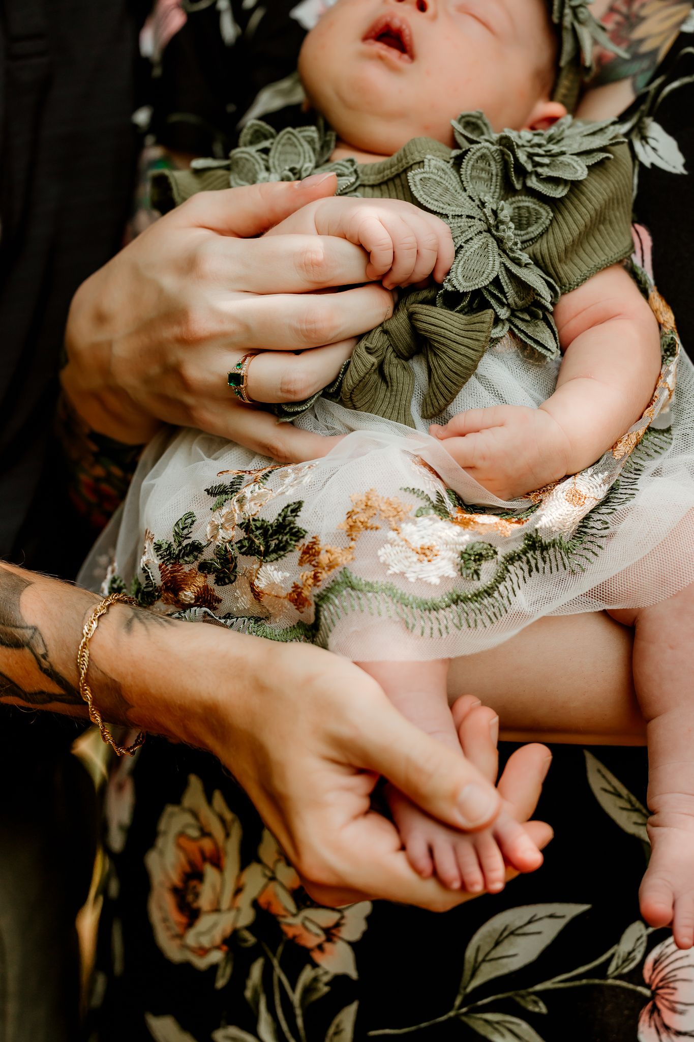 A newborn baby in a green floral dress is cradled in adult hands, one with a tattoo and gold bracelet.