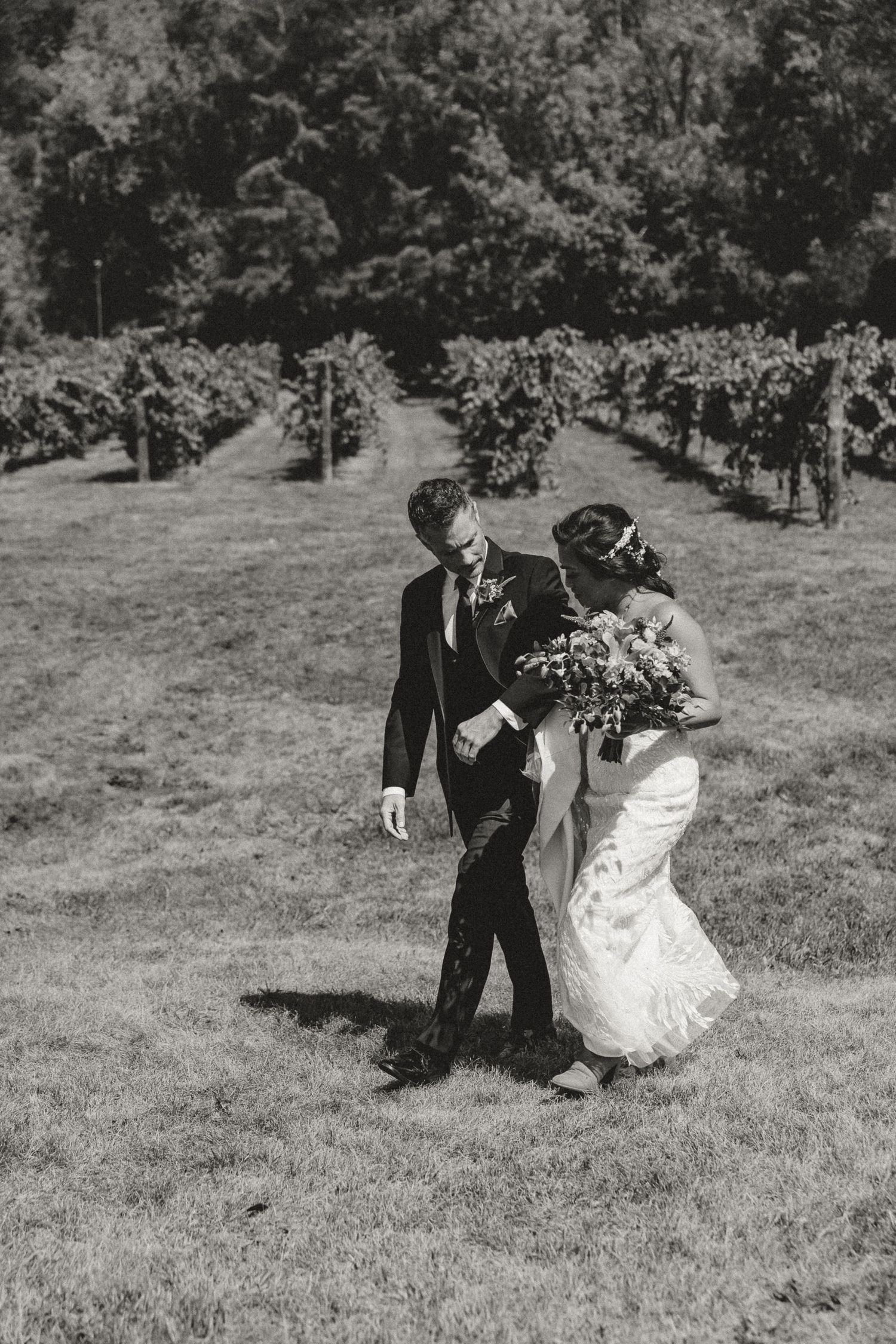 Black and white photo of a bride and groom walking through a vineyard, bride holding a bouquet.
