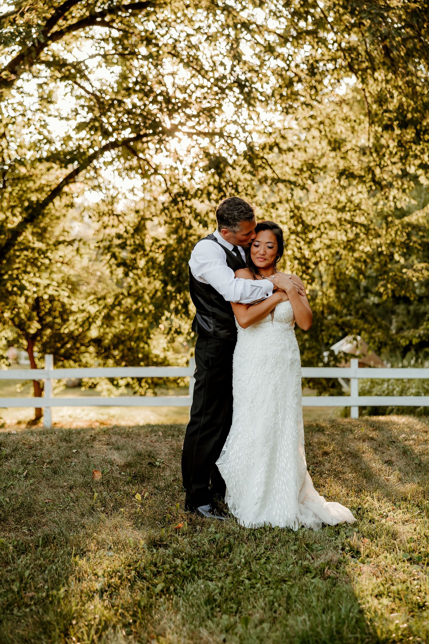 Bride and groom embrace tenderly outdoors near a white fence, surrounded by golden sunlit trees on their wedding day.