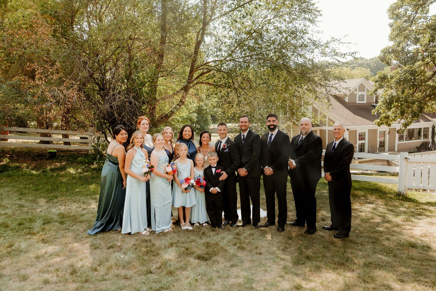 Wedding party posing outdoors on green lawn with bridesmaids in blue gowns and groomsmen in black suits.