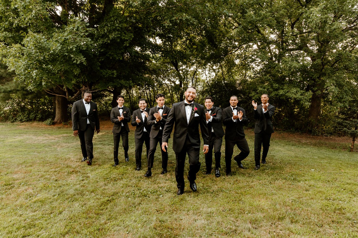 Groom and groomsmen in black tuxedos celebrating outdoors on green grass with trees in background.