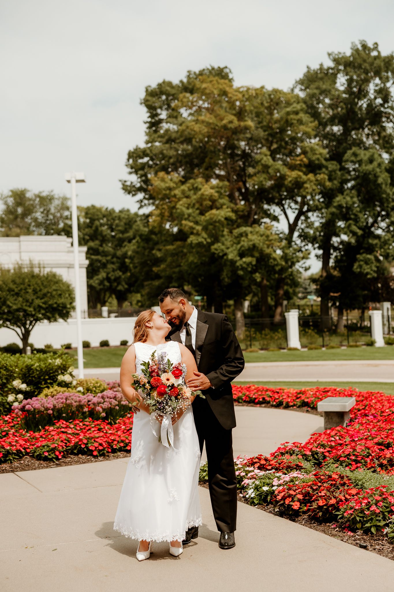 Bride and groom share a kiss outdoors surrounded by vibrant red flowers and lush green trees on their wedding day.