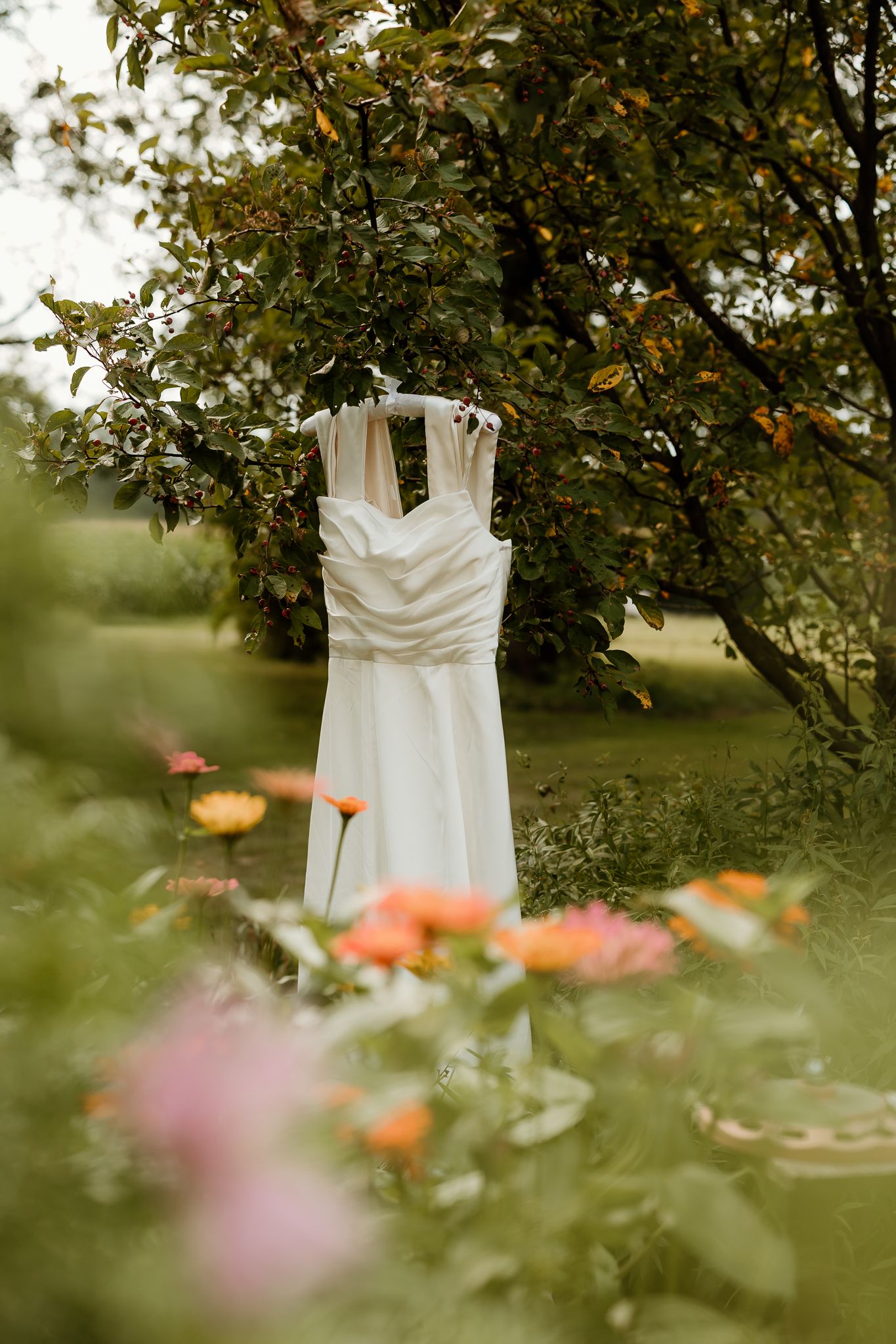 White wedding dress hanging on a tree branch surrounded by colorful garden flowers in a lush green outdoor setting.