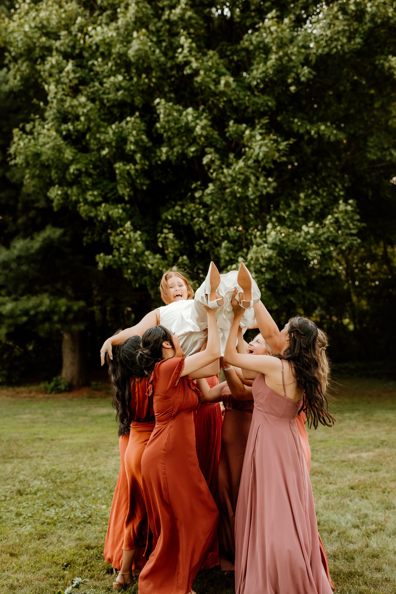 Bridesmaids in rust and pink gowns joyfully toss bride in white dress outdoors near lush green trees.