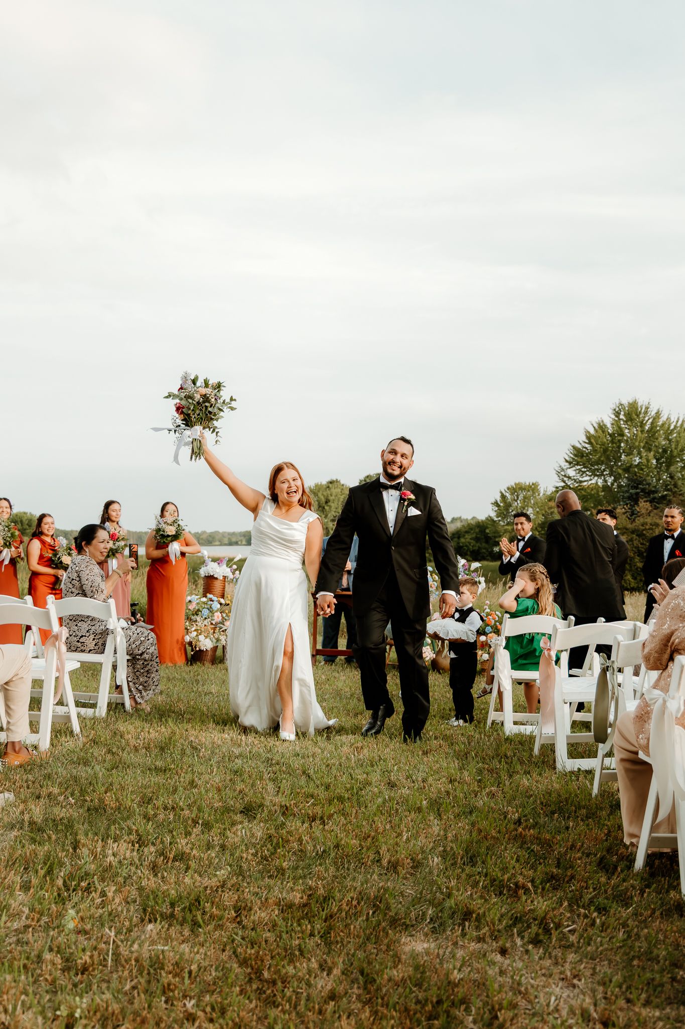 Bride raises bouquet while walking down aisle with groom after outdoor wedding ceremony on green lawn.