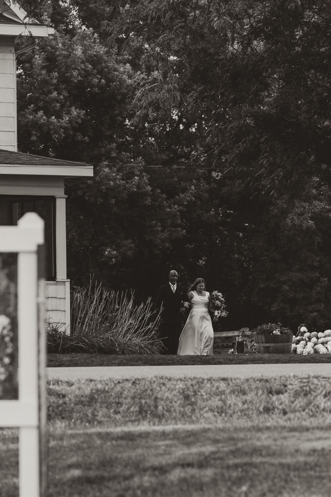 Black and white photo of a bride and groom walking together outdoors near a house surrounded by lush trees.