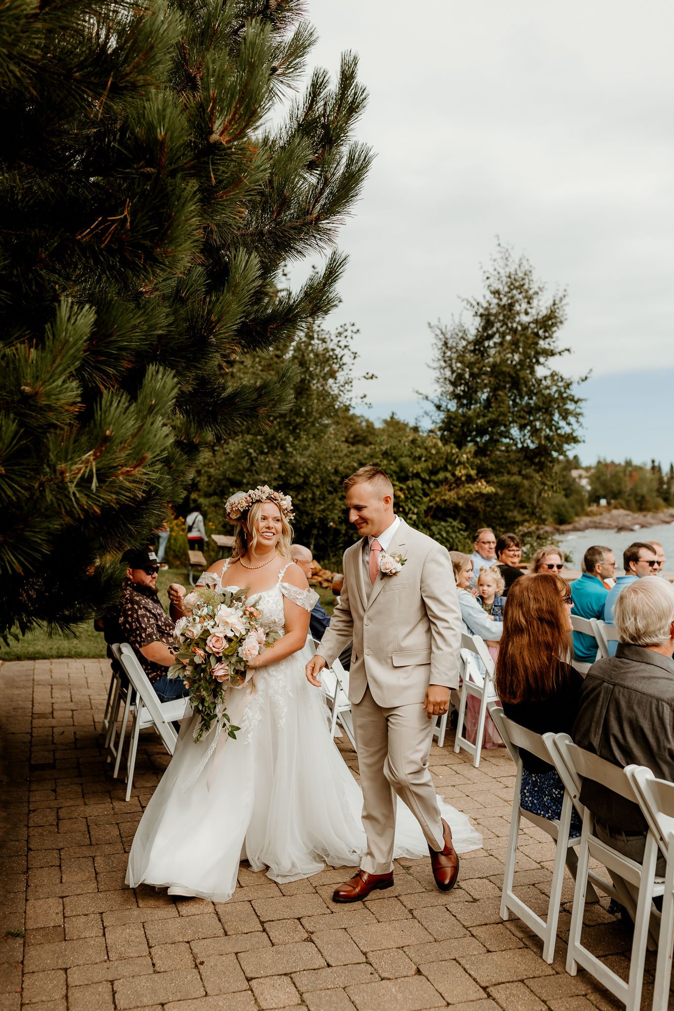 Bride in floral crown and groom in tan suit walk joyfully down outdoor wedding aisle surrounded by seated guests and pine trees.
