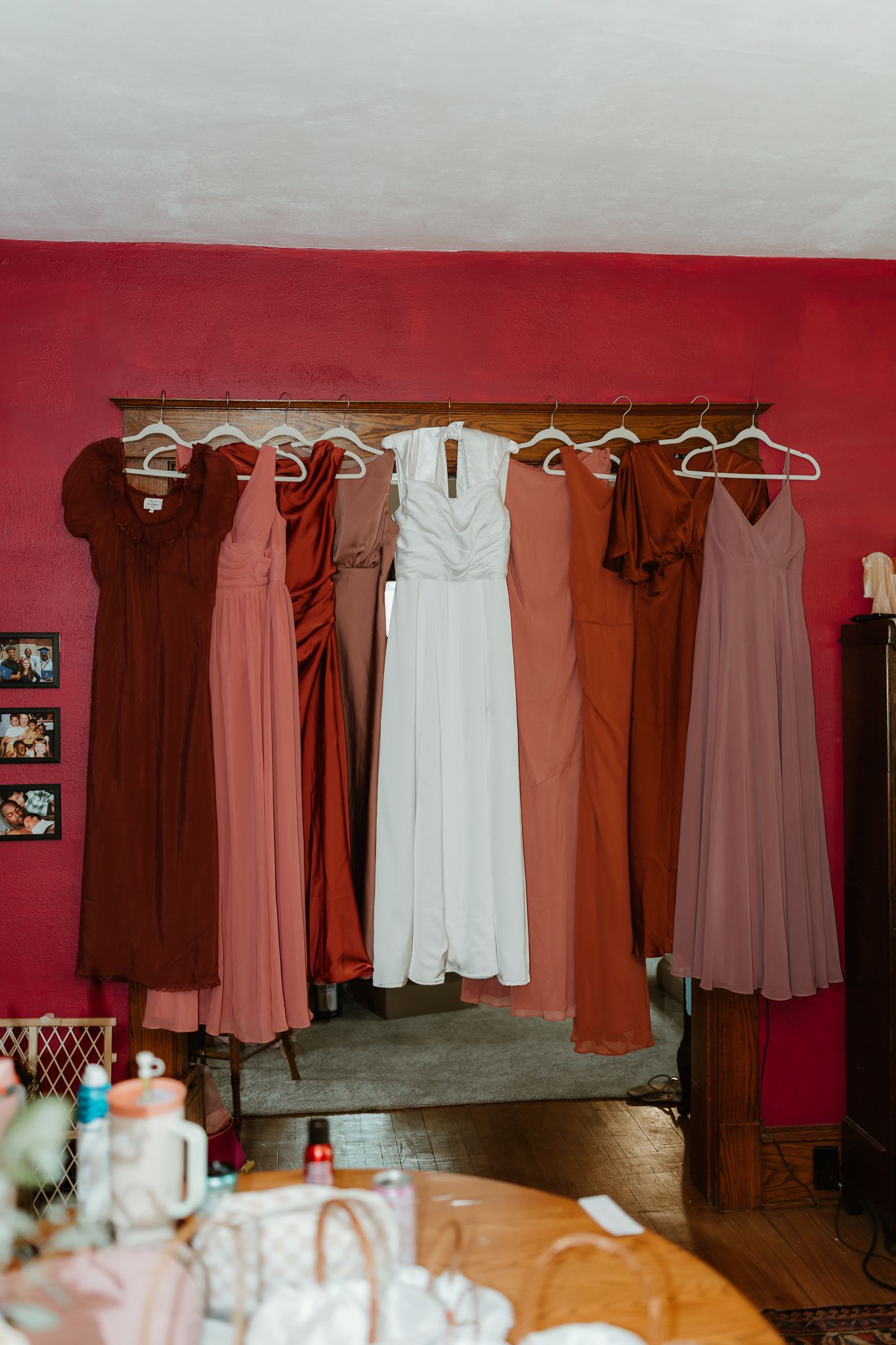 Bridesmaid dresses in shades of rust, mauve, and pink hang alongside a white wedding dress on a wooden rack.