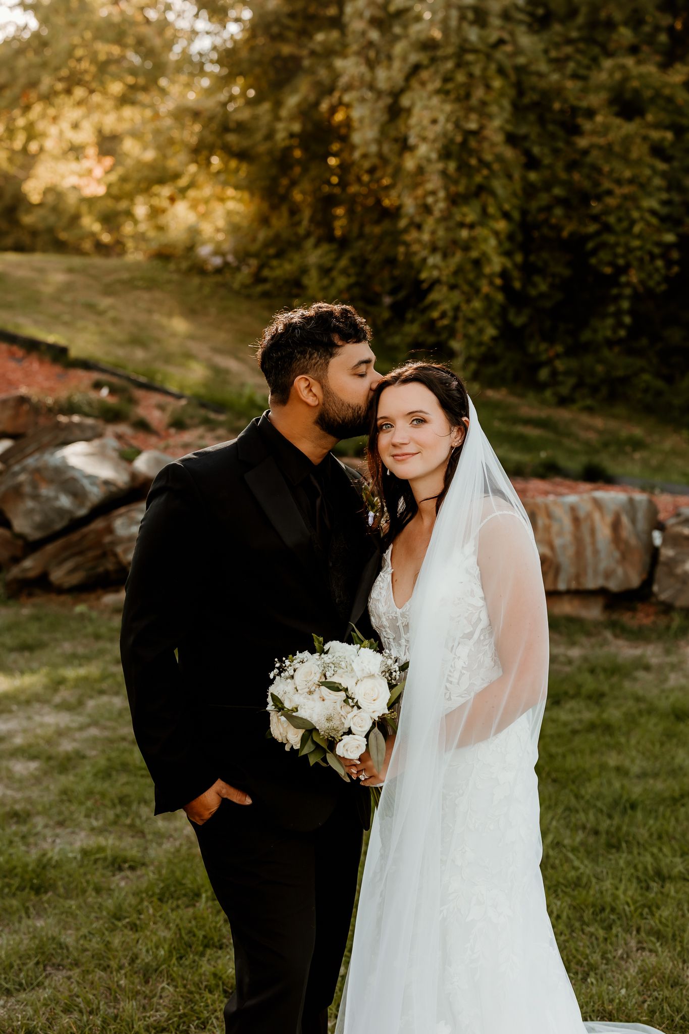 Groom in a black suit kisses the forehead of a smiling bride in a lace gown and veil holding a white rose bouquet outdoors.