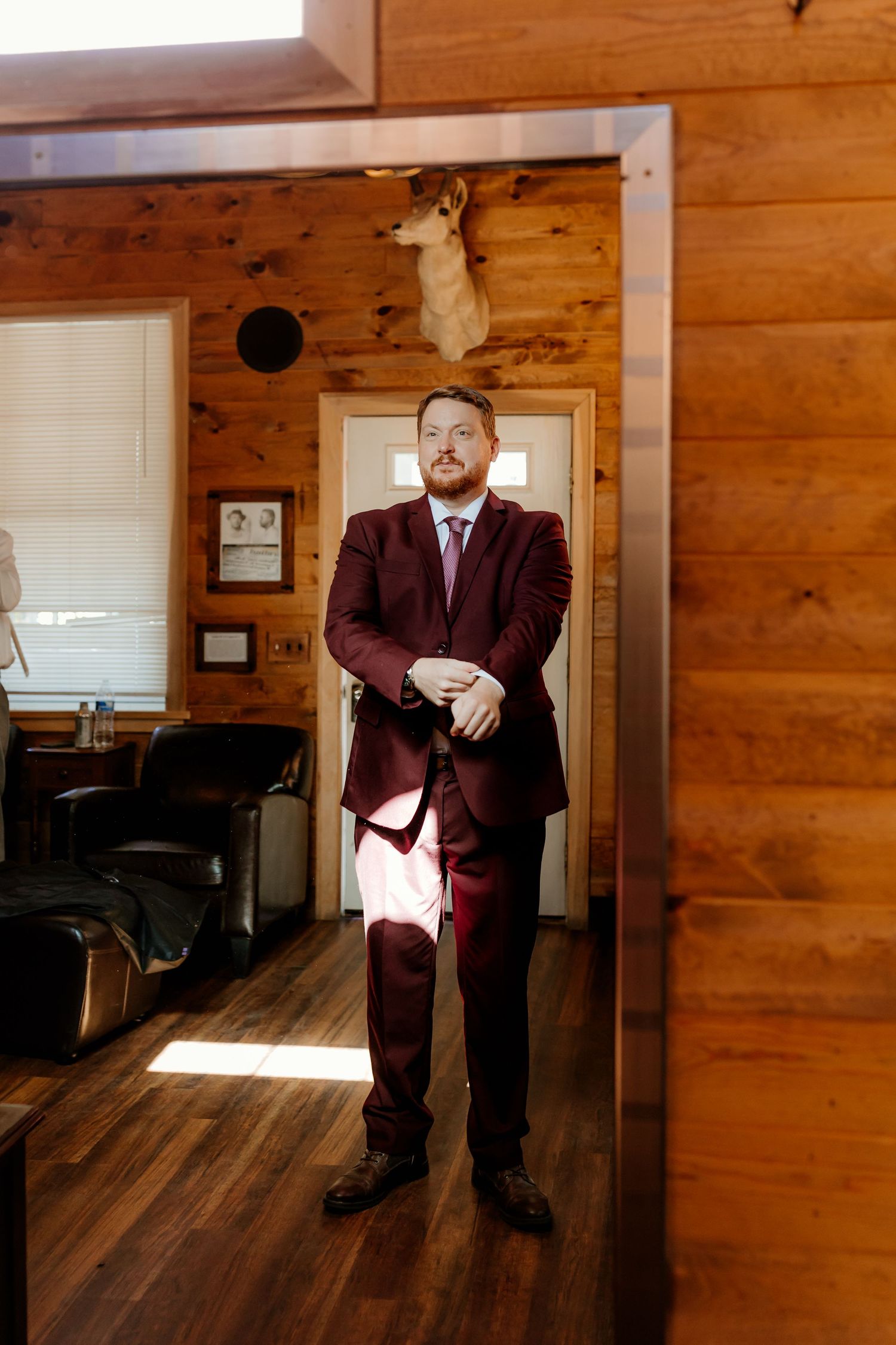 Groom in a burgundy suit and tie adjusts his cuffs while standing in a rustic wood-paneled cabin room with a mounted deer head.