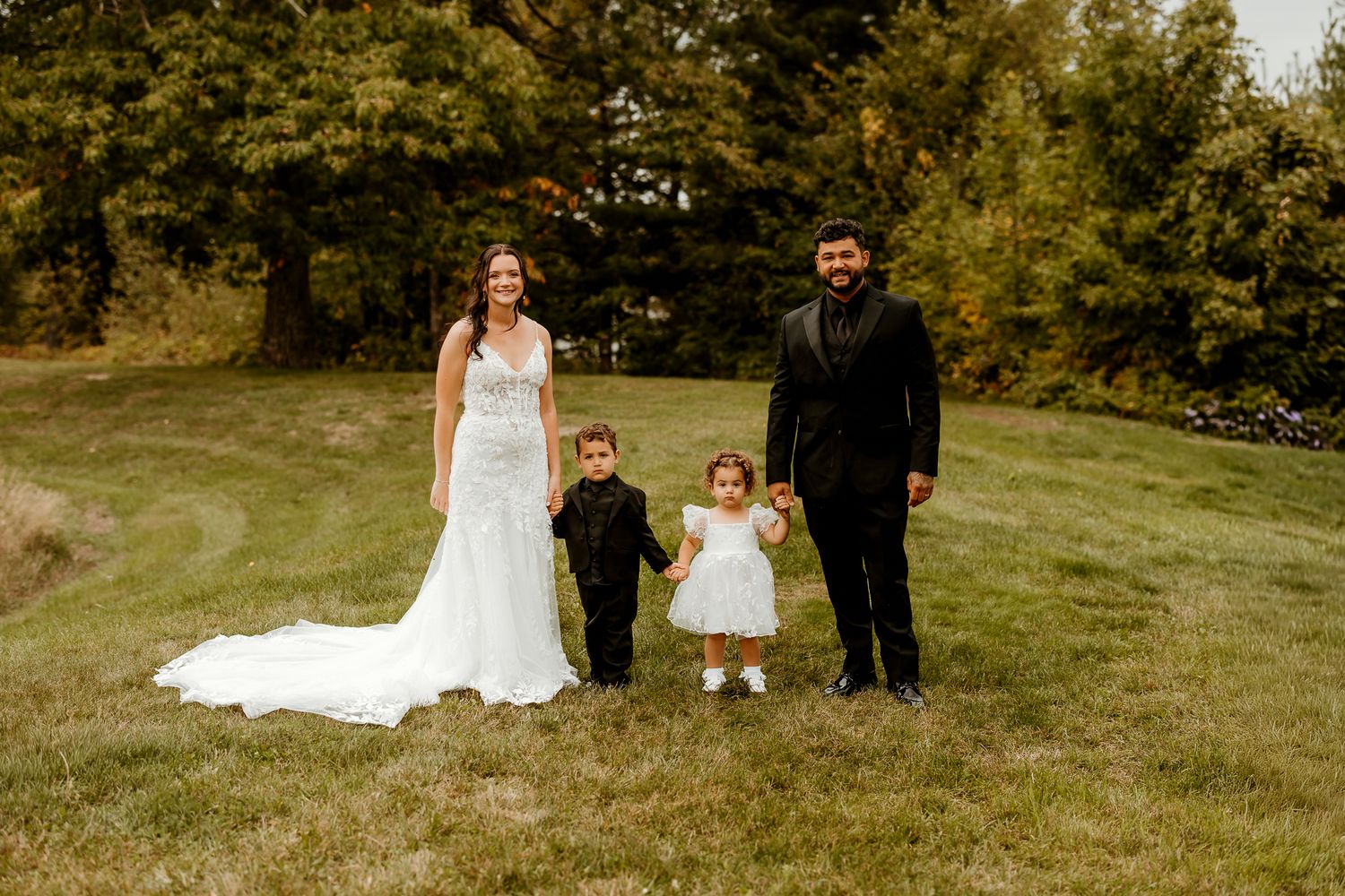 Bride in a white gown and groom in a dark suit hold hands with a young boy and toddler girl on a grassy hillside surrounded by trees.