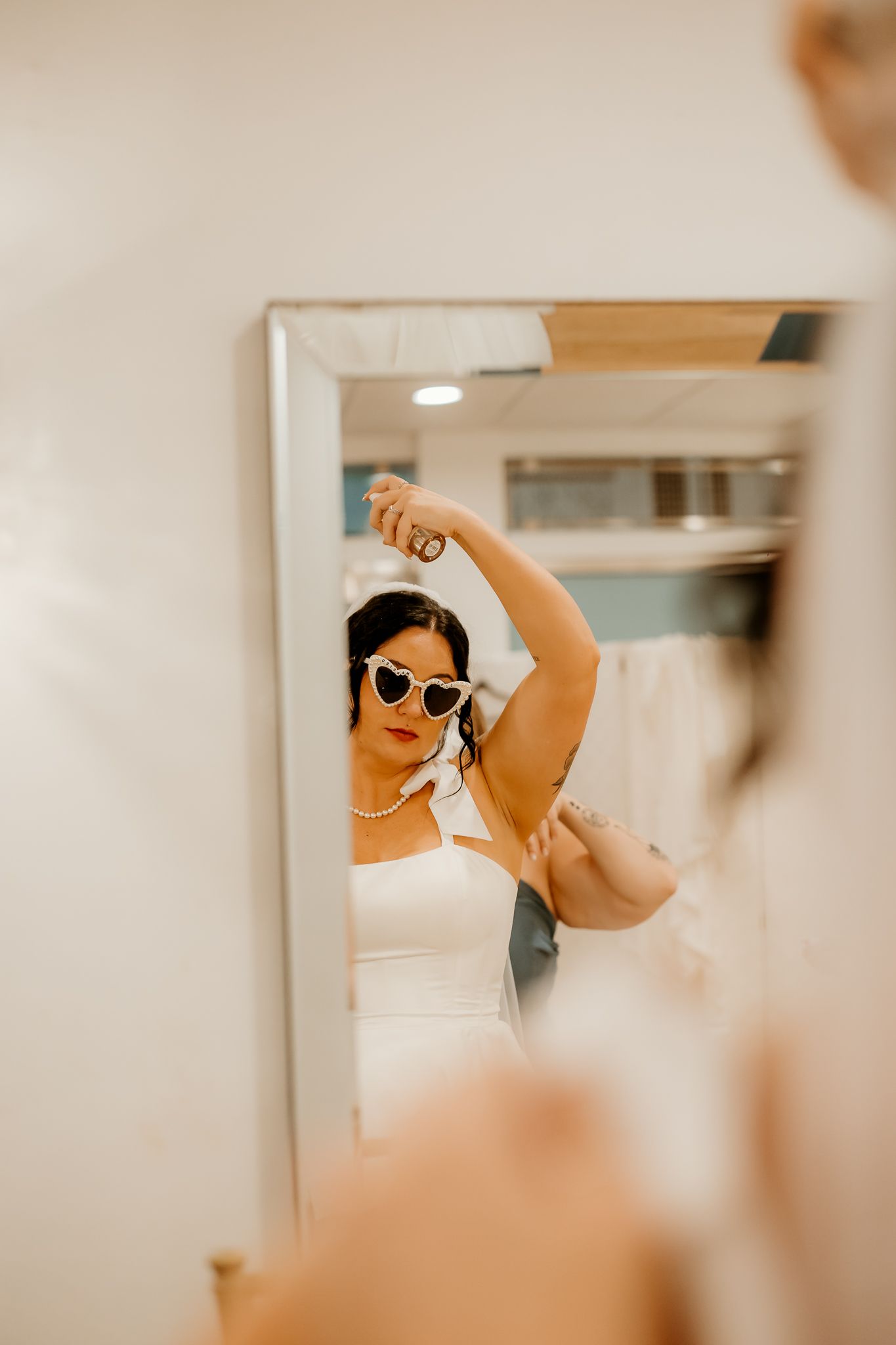 Bride in a white dress and heart-shaped sunglasses spraying her hair in a mirror reflection inside a bridal boutique.