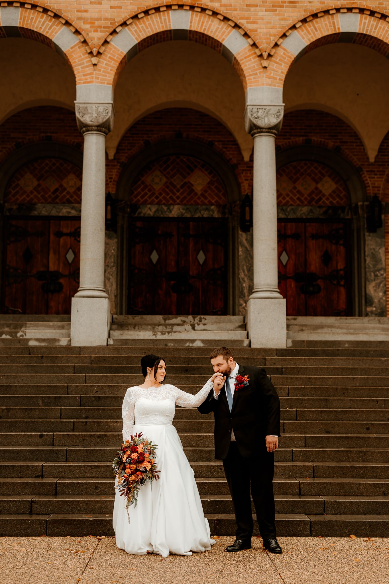 Bride in a white lace gown holding an autumn bouquet as the groom in a black suit kisses her hand on brick church steps.