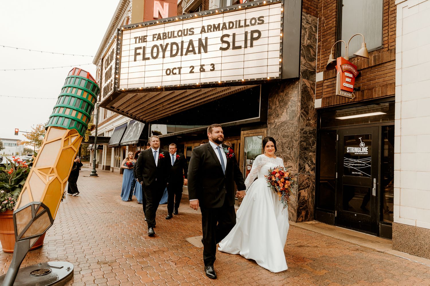 Bride and groom walk hand in hand down a brick sidewalk past a vintage theater marquee, followed by their wedding party.
