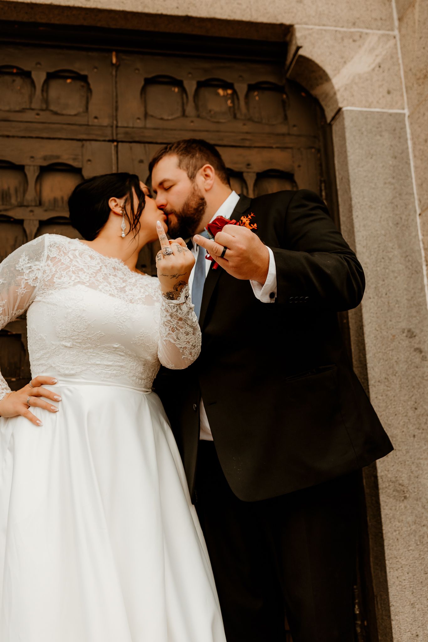 Bride in a white lace-sleeved gown and groom in a dark suit with a boutonniere kiss while showing their rings in front of a stone doorway.