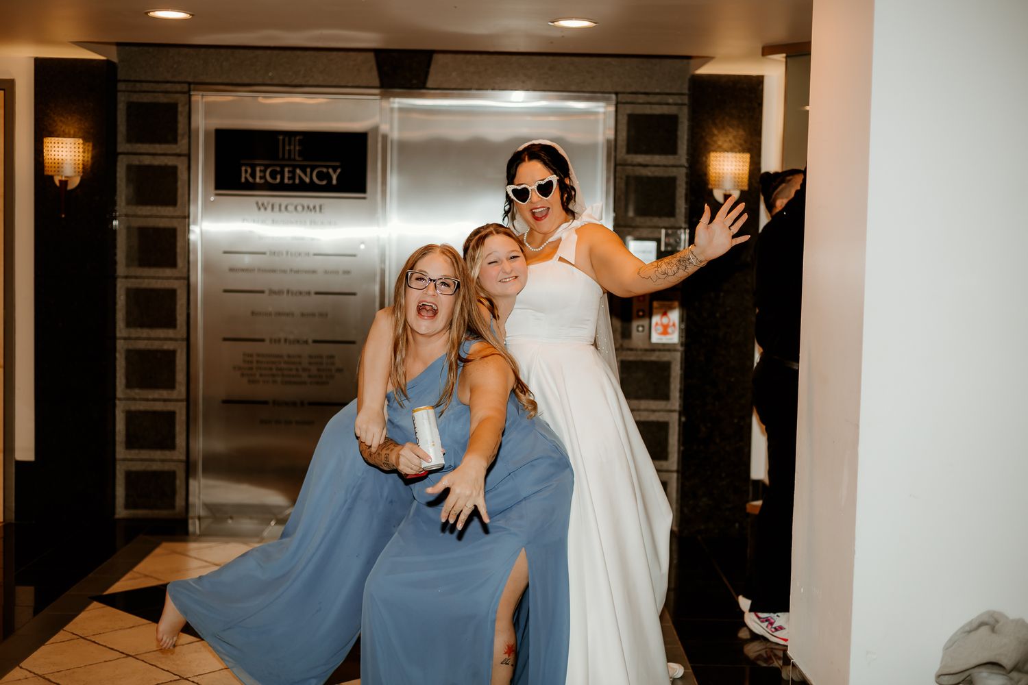 Bride in a white gown and sunglasses poses playfully with two bridesmaids in blue dresses near elevator doors in a hotel lobby.