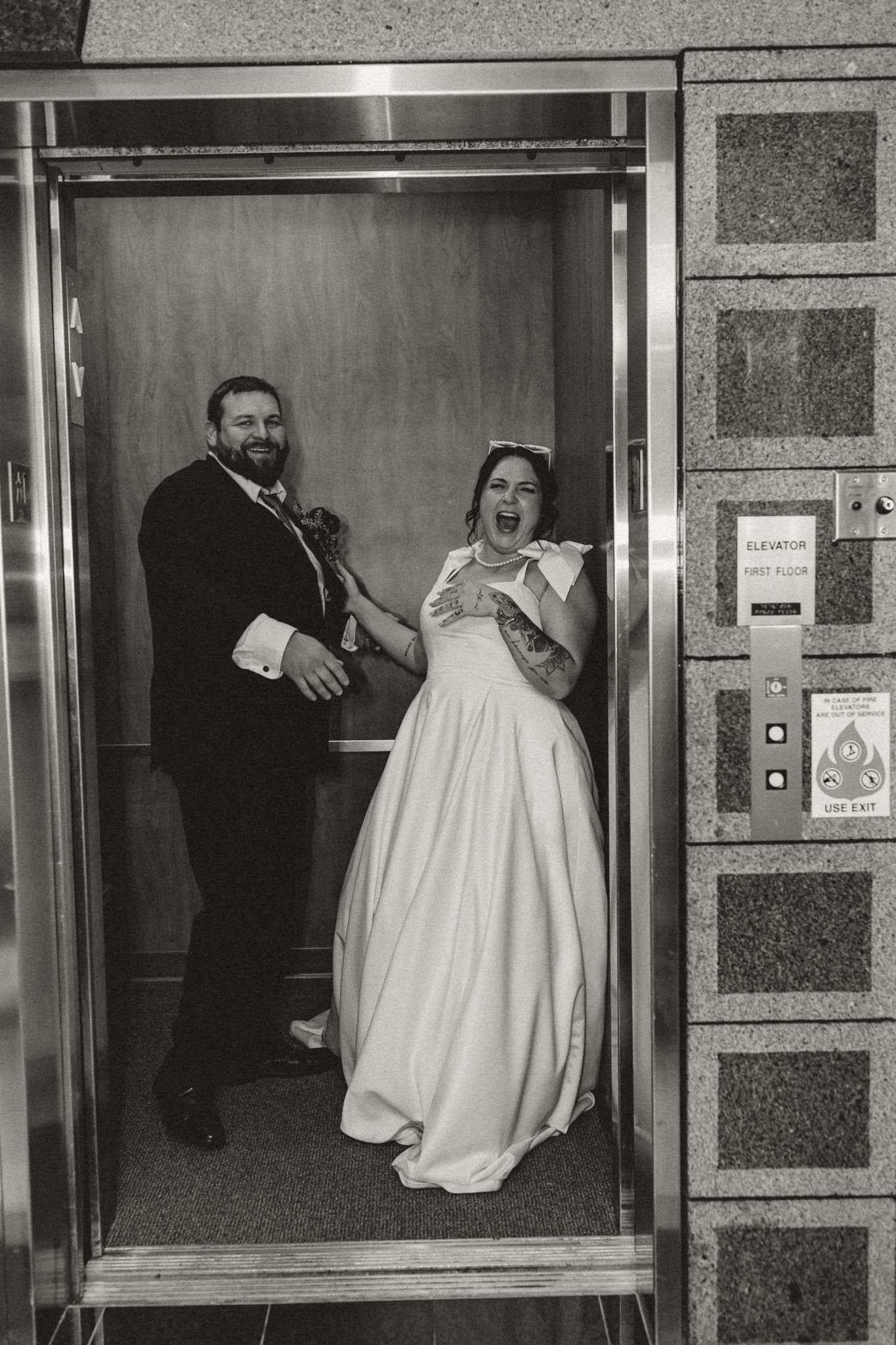 Black and white photo of a laughing bride in a white gown and a smiling groom in a dark suit posing inside a wedding venue elevator.