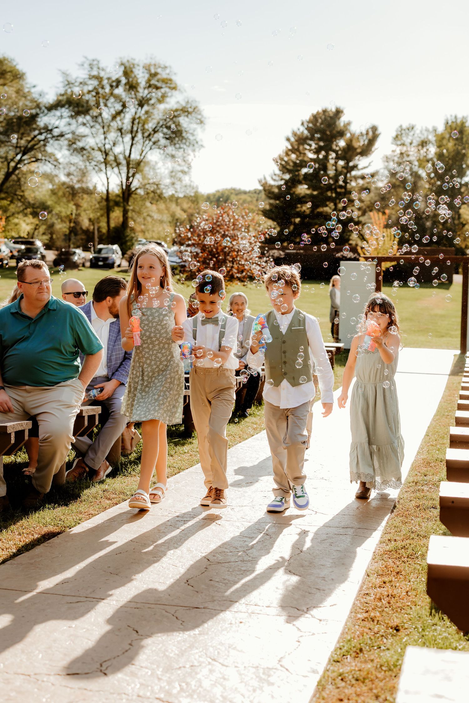 Children in formal attire walk down an outdoor wedding aisle blowing bubbles while guests watch from seats on a sunny day.