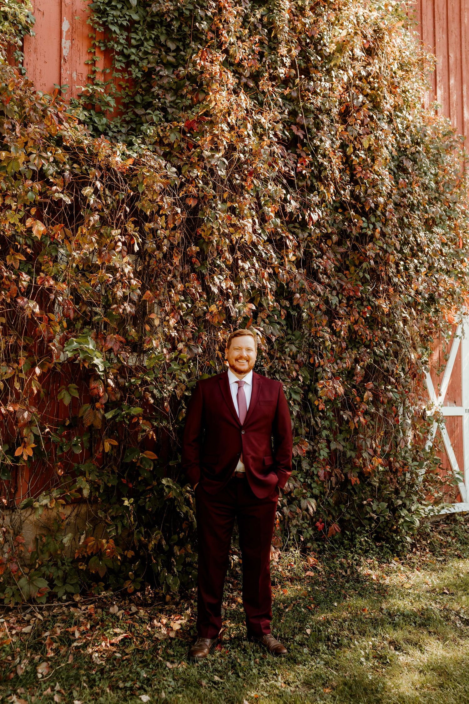 Smiling bearded man in a burgundy suit and tie stands in front of a red barn covered with autumn ivy in warm sunlight.